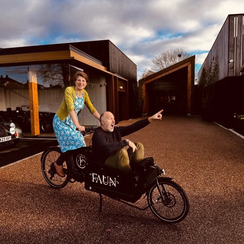 A woman in a blue dress and green cardigan riding a large three-wheeled bike with a man in a black sweater sitting in the front. The man is pointing and smiling, while the woman is smiling. They are outside in a modern urban area with dark buildings and a parking lot, and the sky is partly cloudy.