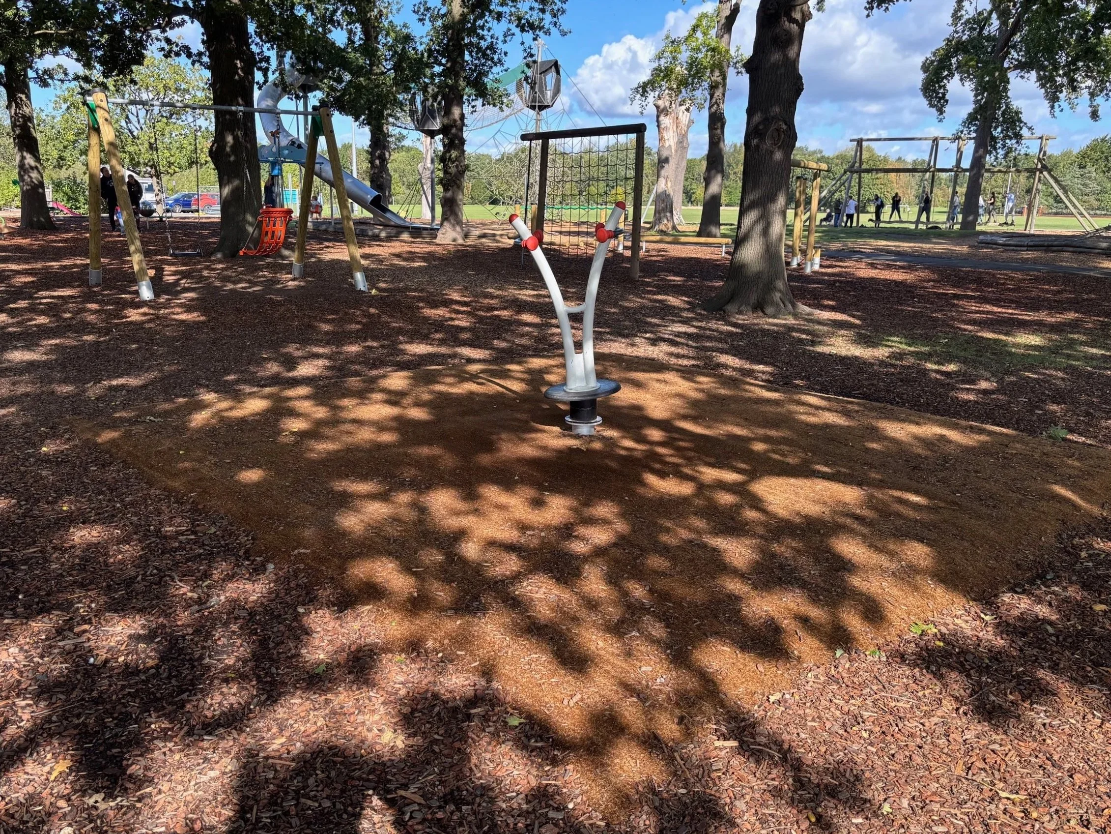 Children playing on outdoor playground equipment including a slide, swings, and climbing structures, surrounded by trees and a grassy field under a partly cloudy sky.