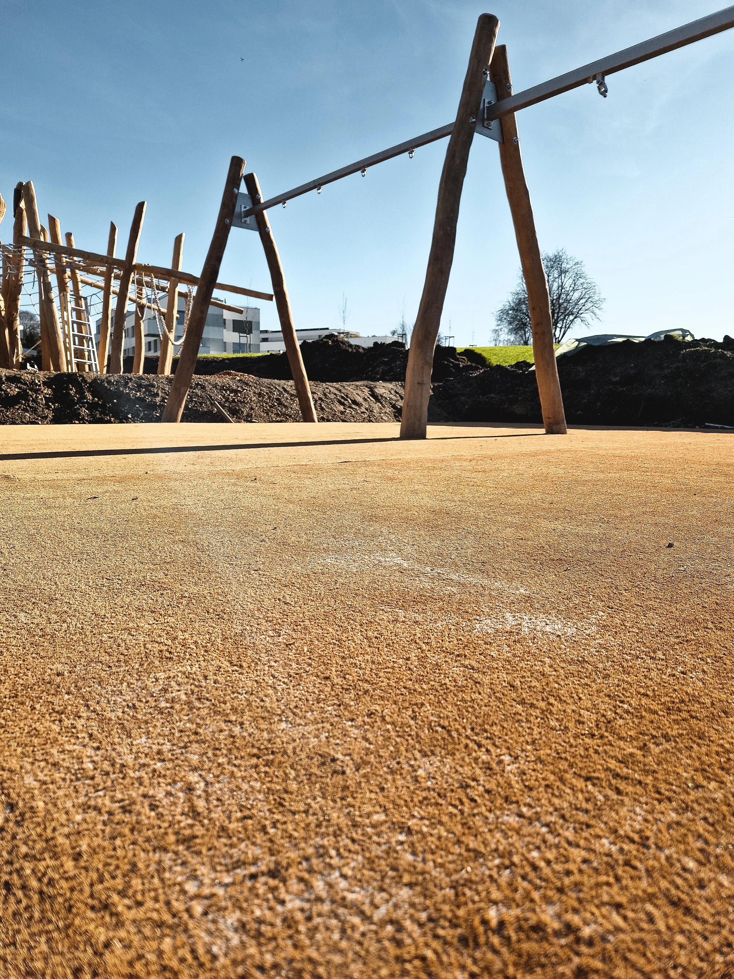 A playground with wooden swings and climbing structures on a sandy surface, with a grassy hill, trees, and buildings in the background under a clear blue sky.