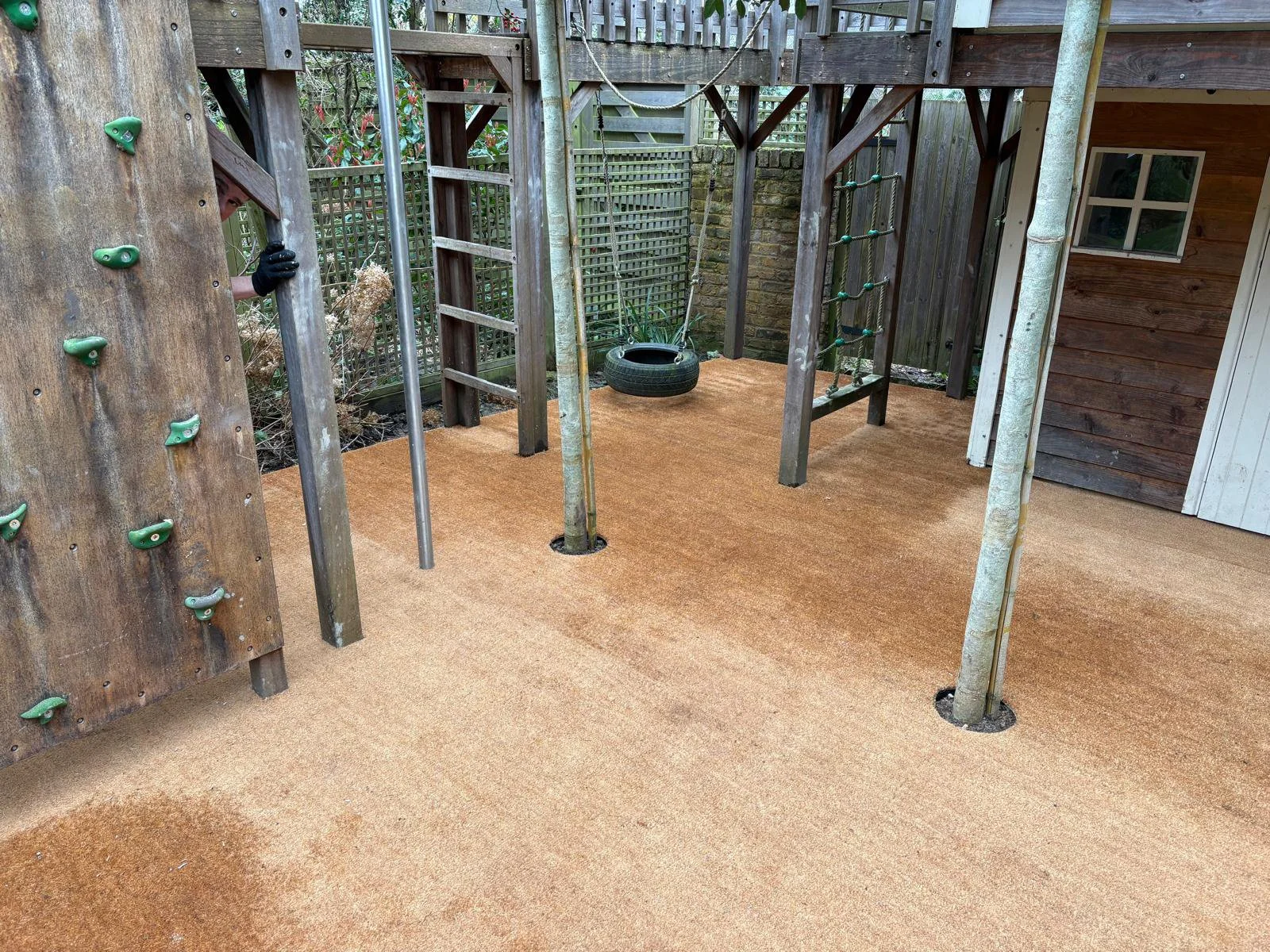 Outdoor play area with a climbing wall, a tire swing, and a wooden play structure with a ladder and ropes, set on a soft orange surface.