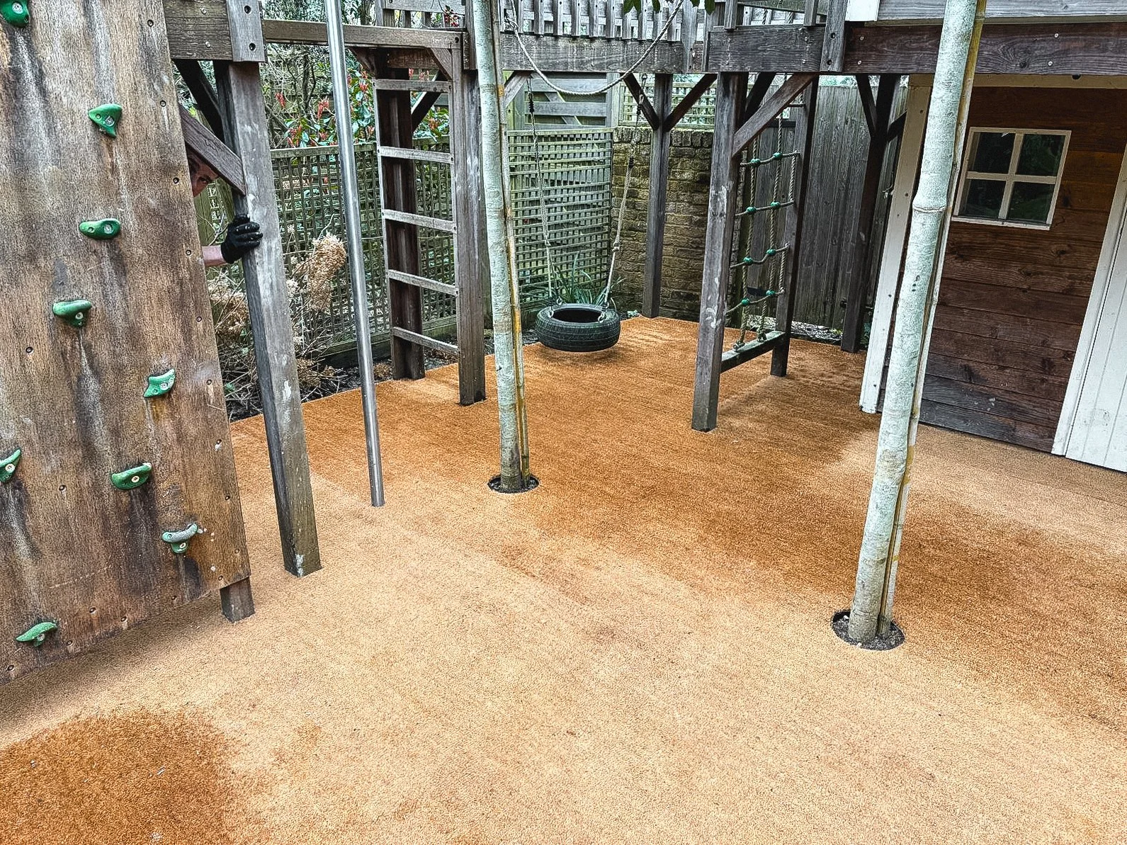 Outdoor play area with a climbing wall, a tire swing, and a wooden play structure with a ladder and ropes, set on a soft orange surface.