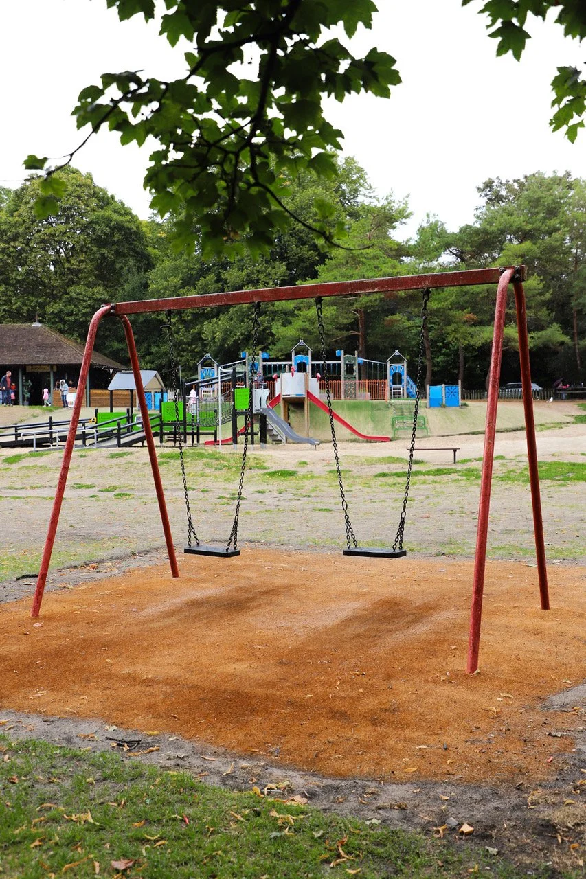 Empty swing set in a park with a playground in the background, surrounded by trees.