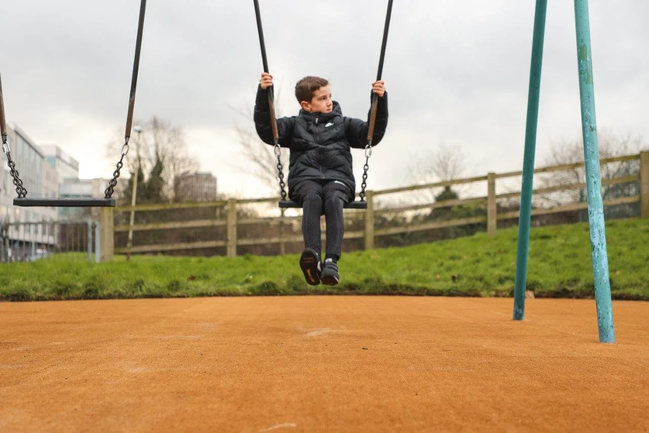 A young boy sitting on a swing at a playground, wearing a black jacket and pants, with a grassy hill and trees in the background.