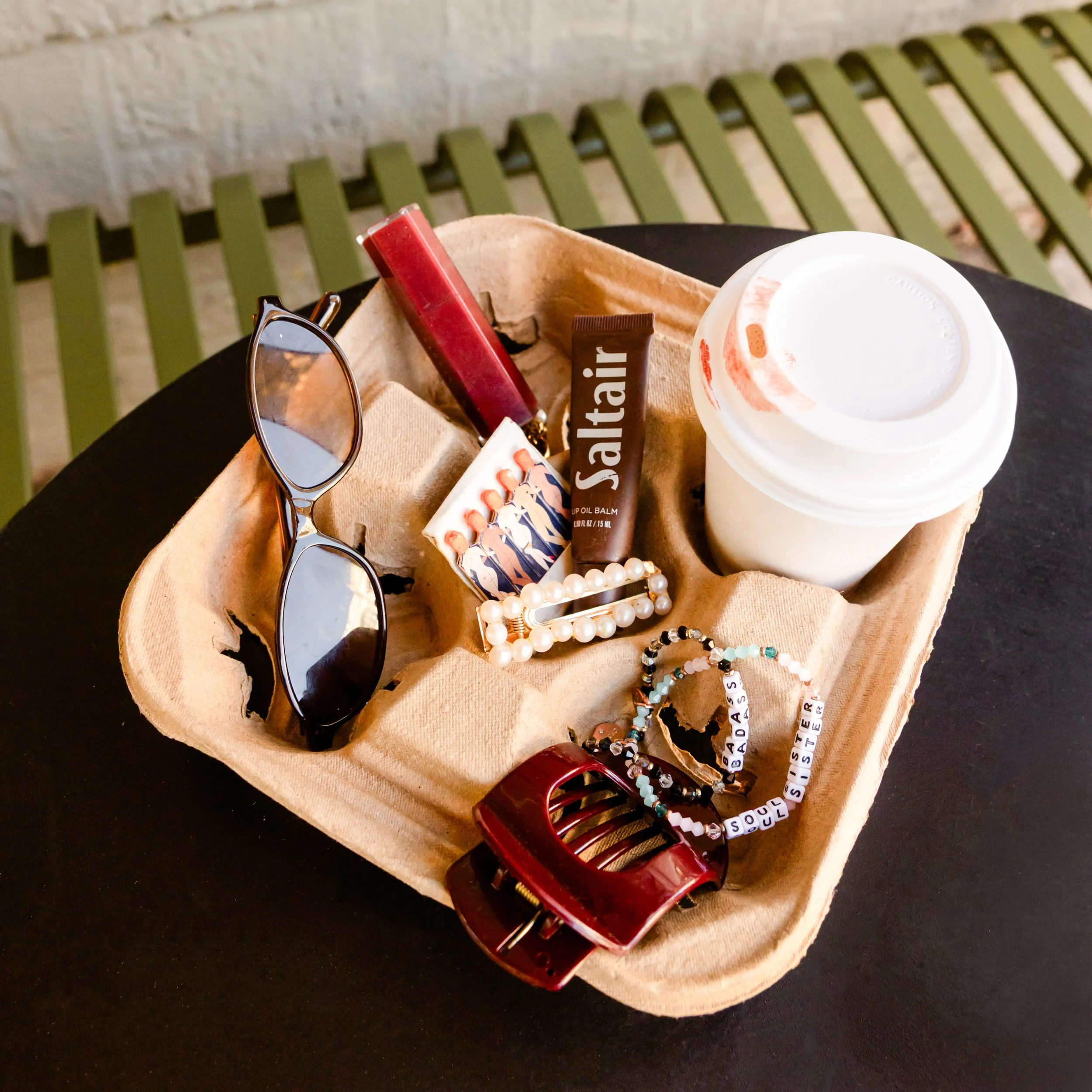 Lifestyle flatlay featuring sunglasses, coffee, lip balm, and accessories styled on a cafe table, representing the Brand Curators creator brand aesthetic.