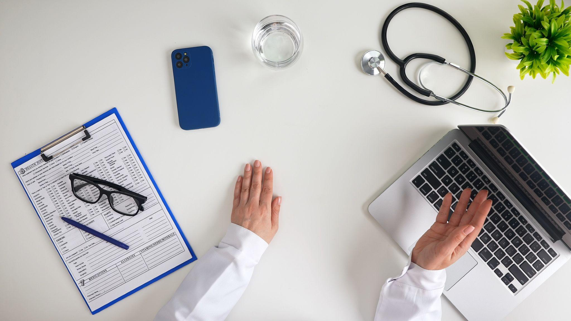desk with medical items including a clipboard with a health history form and glasses, a blue smartphone, a glass of water, a stethoscope, a laptop, and a green plant; a person wearing a white coat reaching for the laptop.
