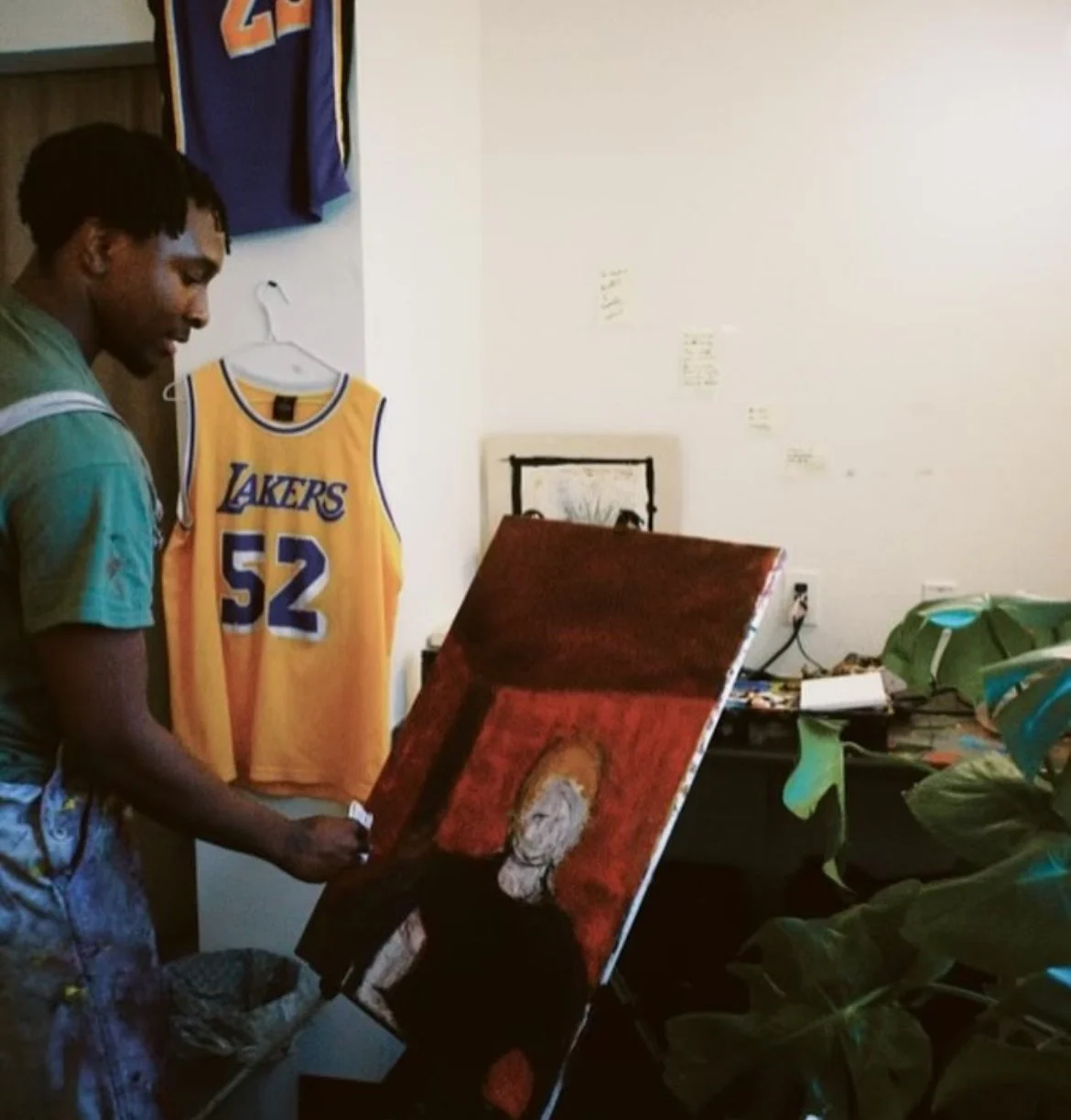A young man painting a portrait in a room with Lakers jerseys hanging on the wall and plants on a desk.