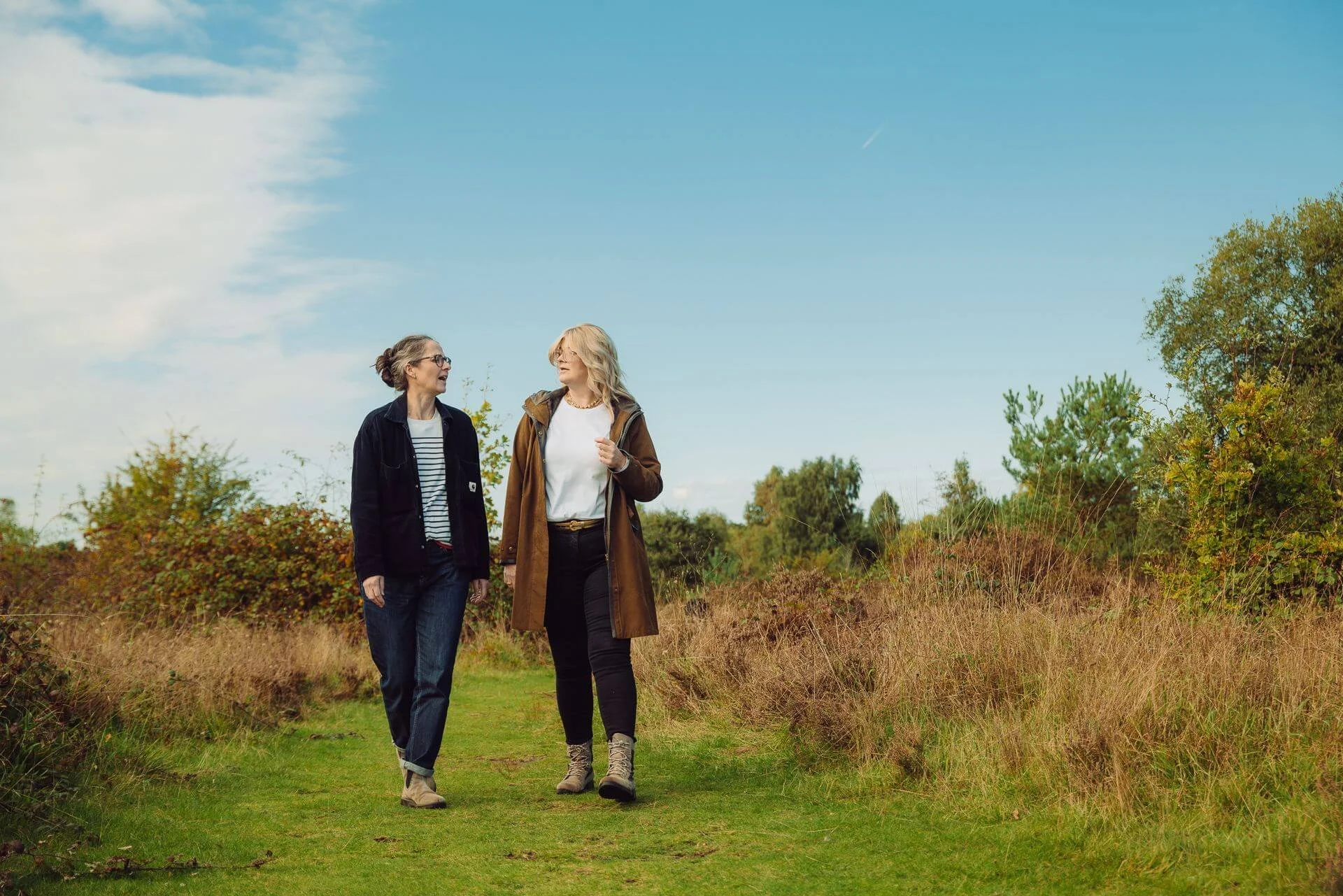 Two women walking and talking outdoors on a grassy trail surrounded by autumn foliage, under a mostly cloudy sky.