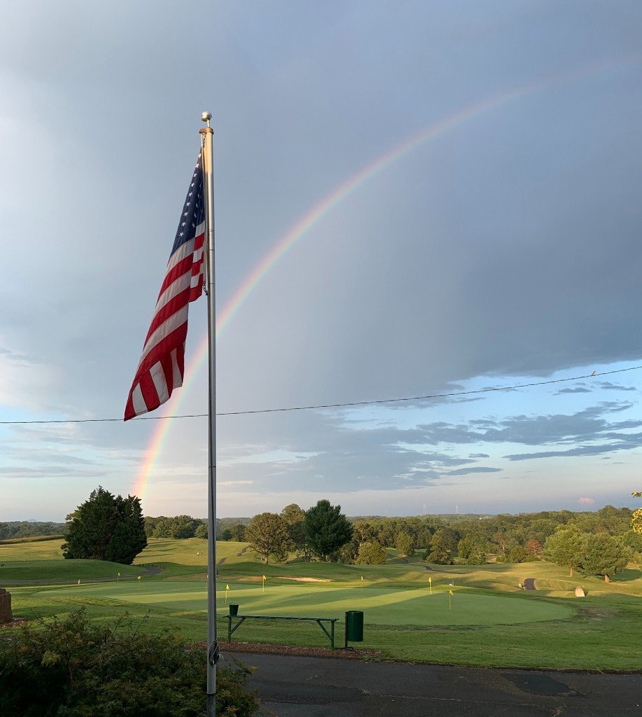 An American flag on a flagpole with a rainbow in the sky above a golf course.