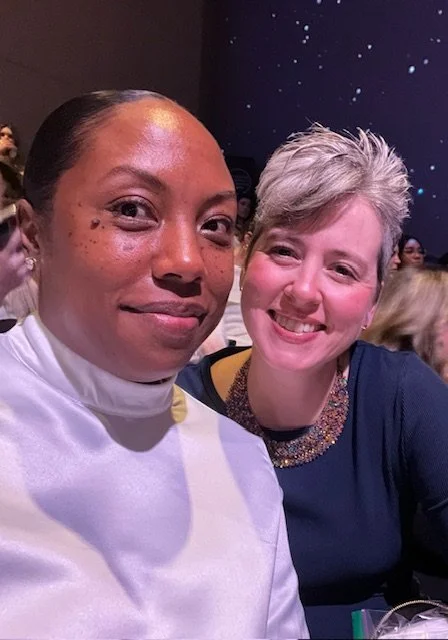 Two women taking a selfie together at an event, one with dark hair and freckles in a white top, and the other with short, gray hair in a dark top with a sparkly neckline, smiling at the camera.