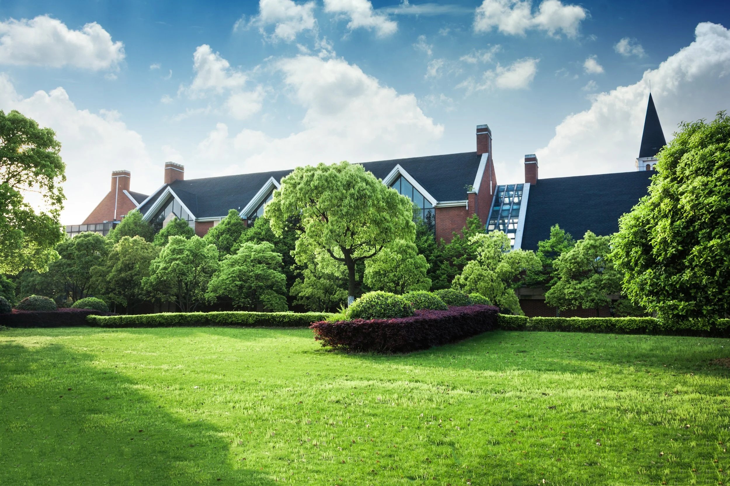 A large, institutional-style building with peaked roofs, brick walls, and a tall steeple in the background, surrounded by lush, green trees and well-maintained lawn under a partly cloudy sky.