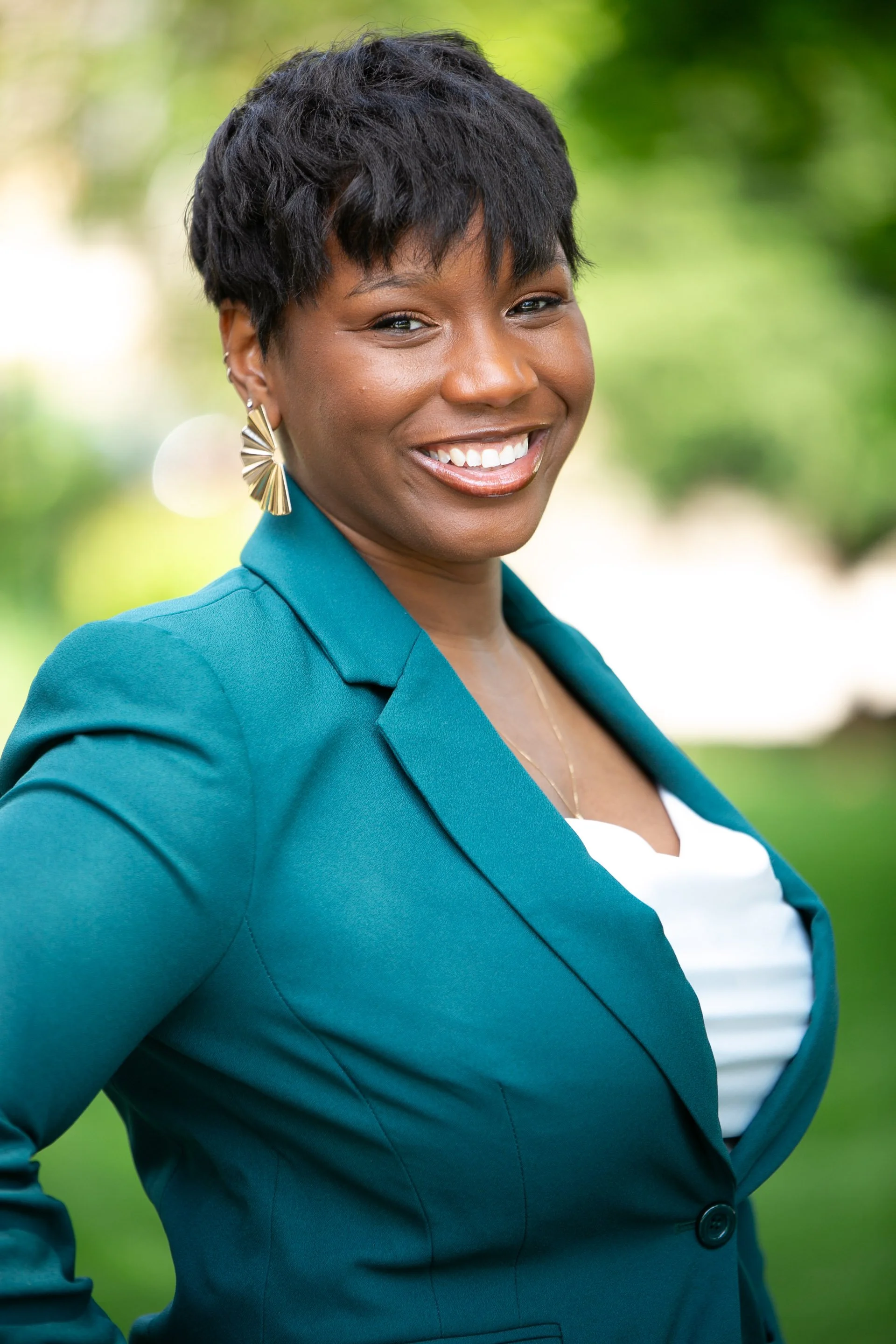 A smiling woman with short black hair, wearing a teal blazer and large gold earrings, standing outdoors with blurred green background.