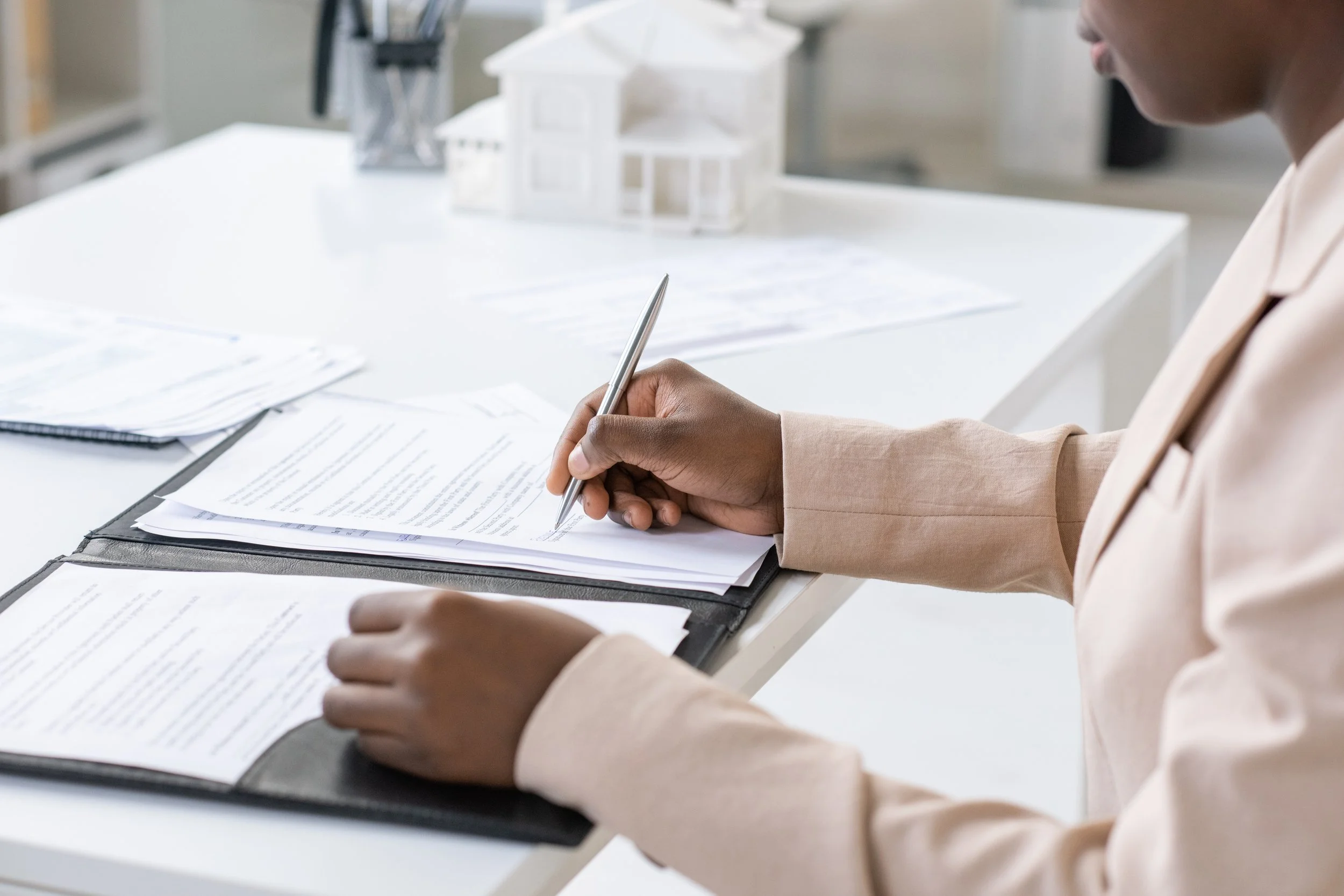 Person in a beige blazer reviewing documents with a pen at a white desk, with a small white model house in the background.
