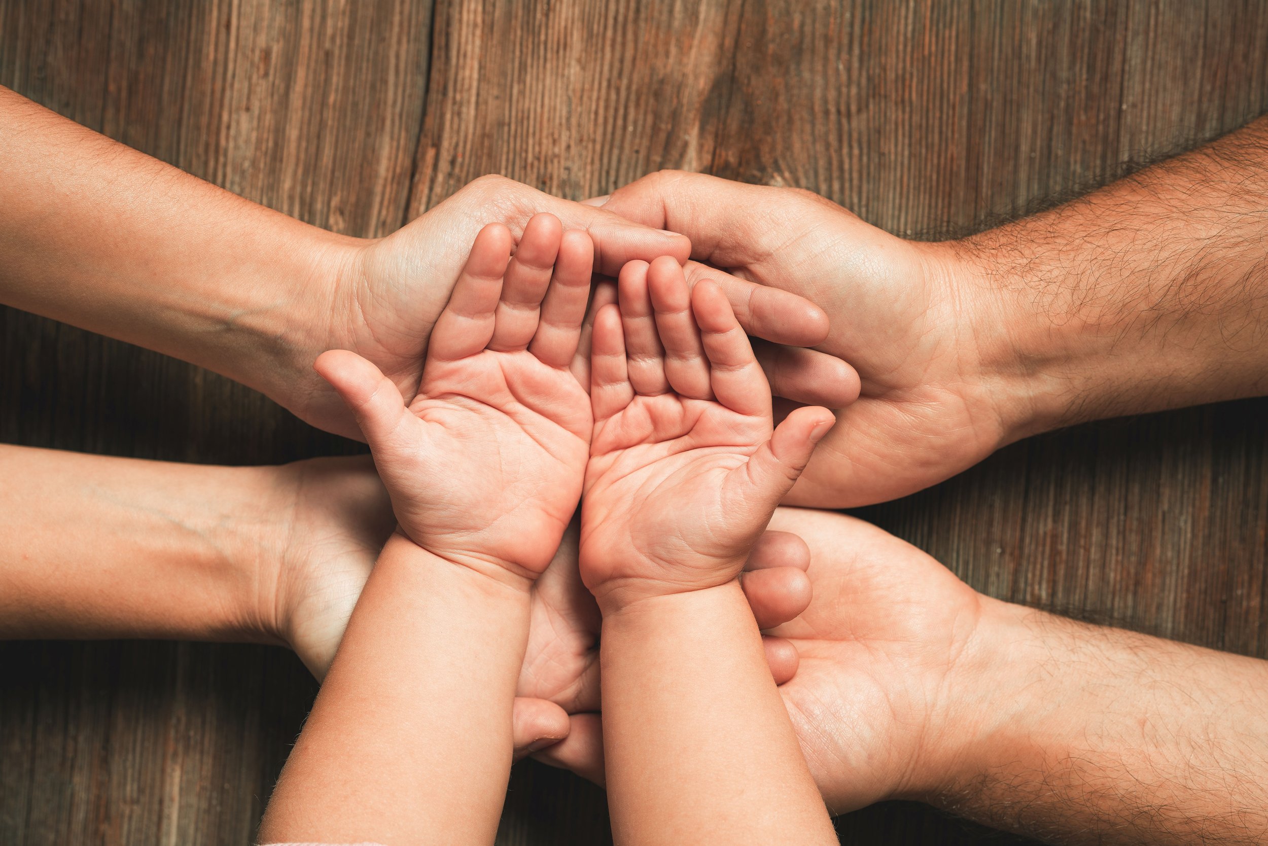 Multiple hands, including children and adults, are stacked together on a wooden surface in a gesture of unity and teamwork.