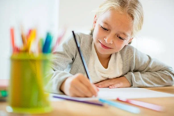 A young girl with blonde hair sits at a table, writing or drawing in a notebook with a pencil, surrounded by colorful markers or pens in a green container.