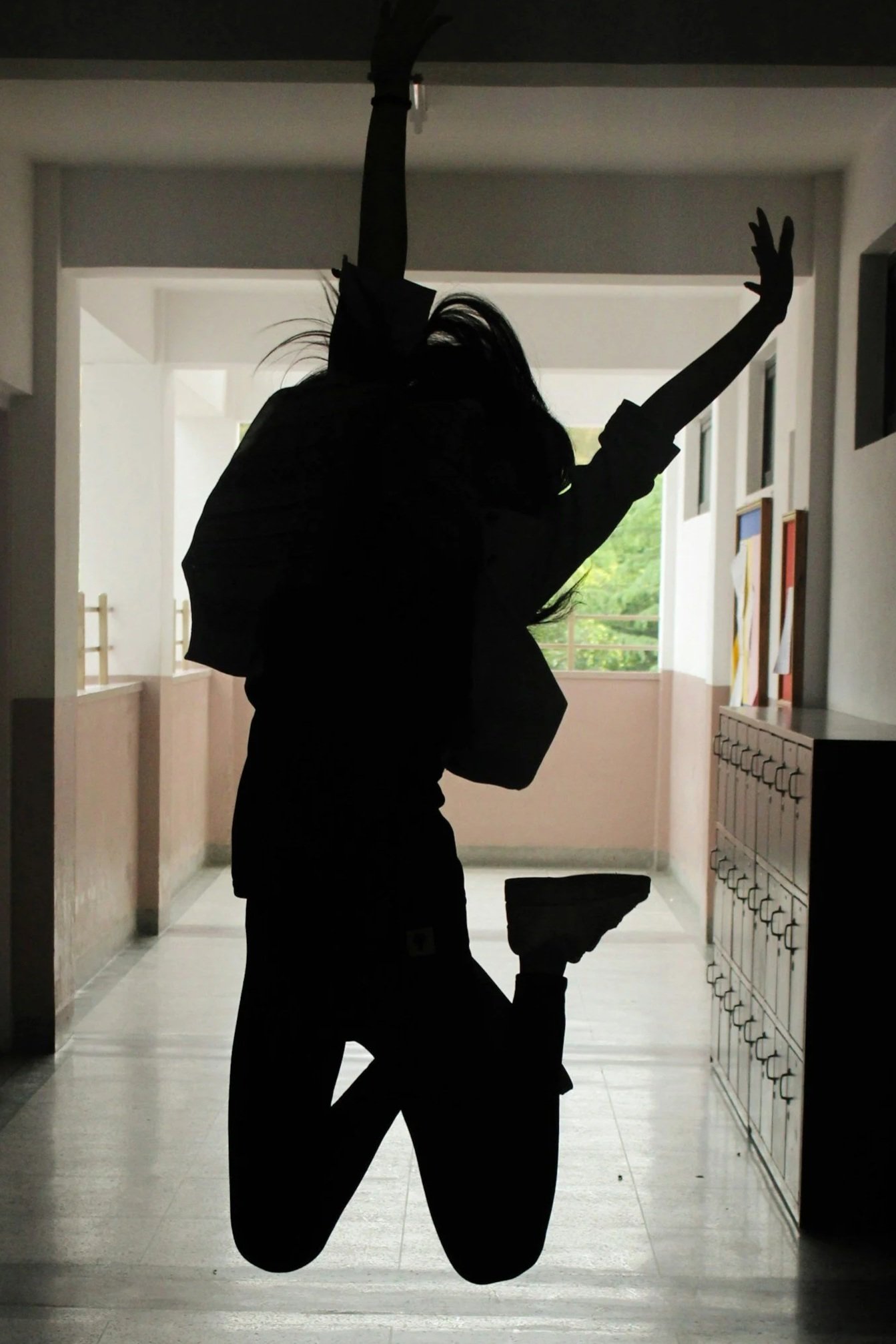 Silhouette of a person kneeling on one knee inside a hallway, with one arm raised and the other arm extending outward, in front of a window with greenery visible outside.