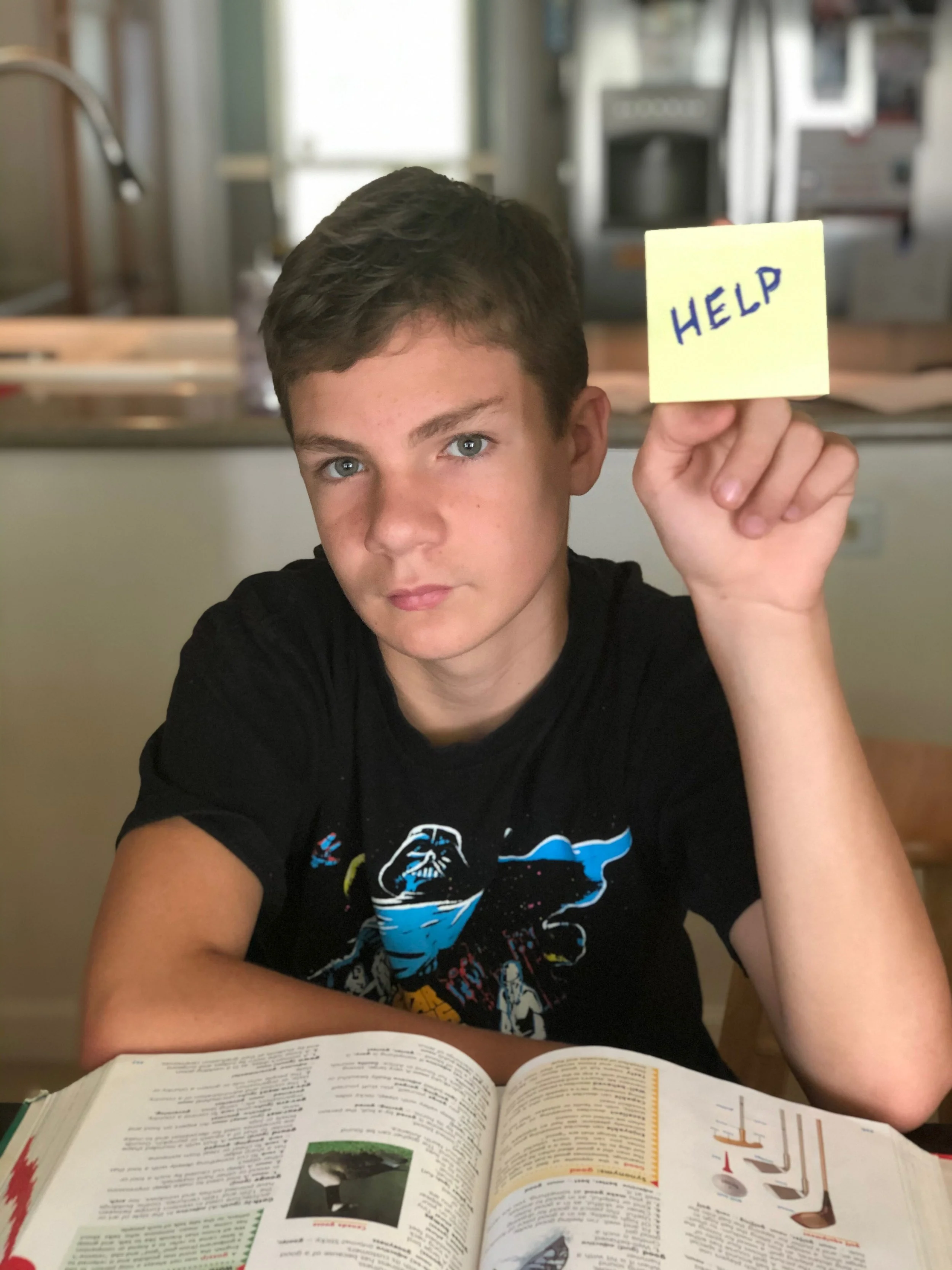 A young boy with short brown hair and blue eyes is sitting at a table with an open book in front of him, holding a yellow sticky note with the word 'HELP' written on it in blue marker.