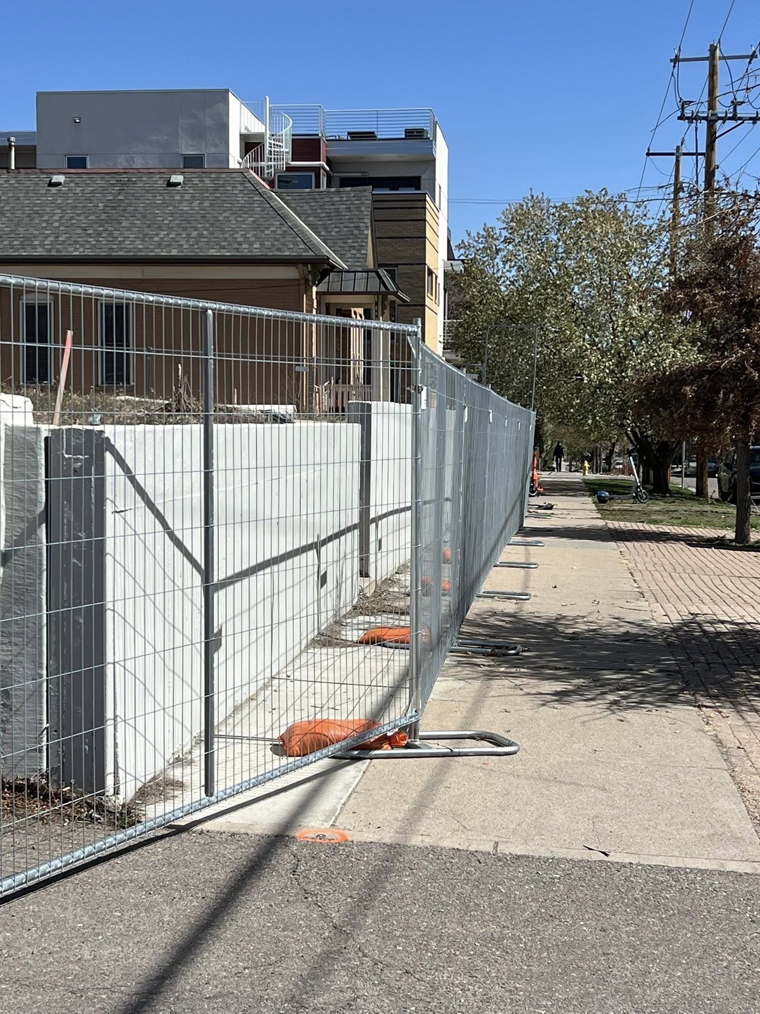 Construction fence along a sidewalk with trees and buildings in the background on a sunny day.