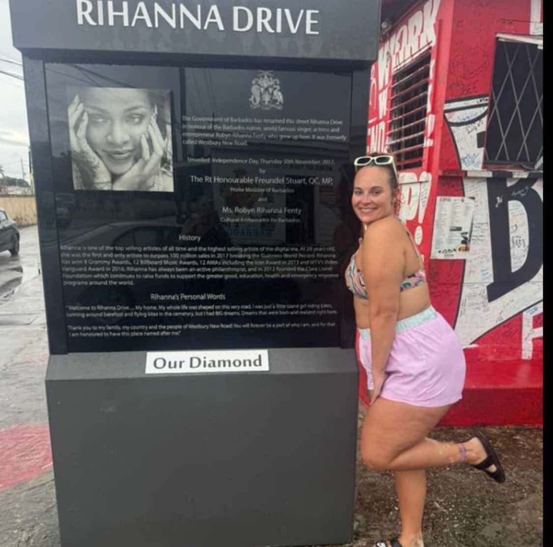 A woman in pink shorts and a colorful top posing next to a black informational display board with a picture of a woman at the top. The display board is titled 'RIHANNA DRIVE' and contains text about Rihanna, her achievements, and personal words. The woman is smiling, wearing sunglasses on her head, and is next to a red graffiti-covered wall.