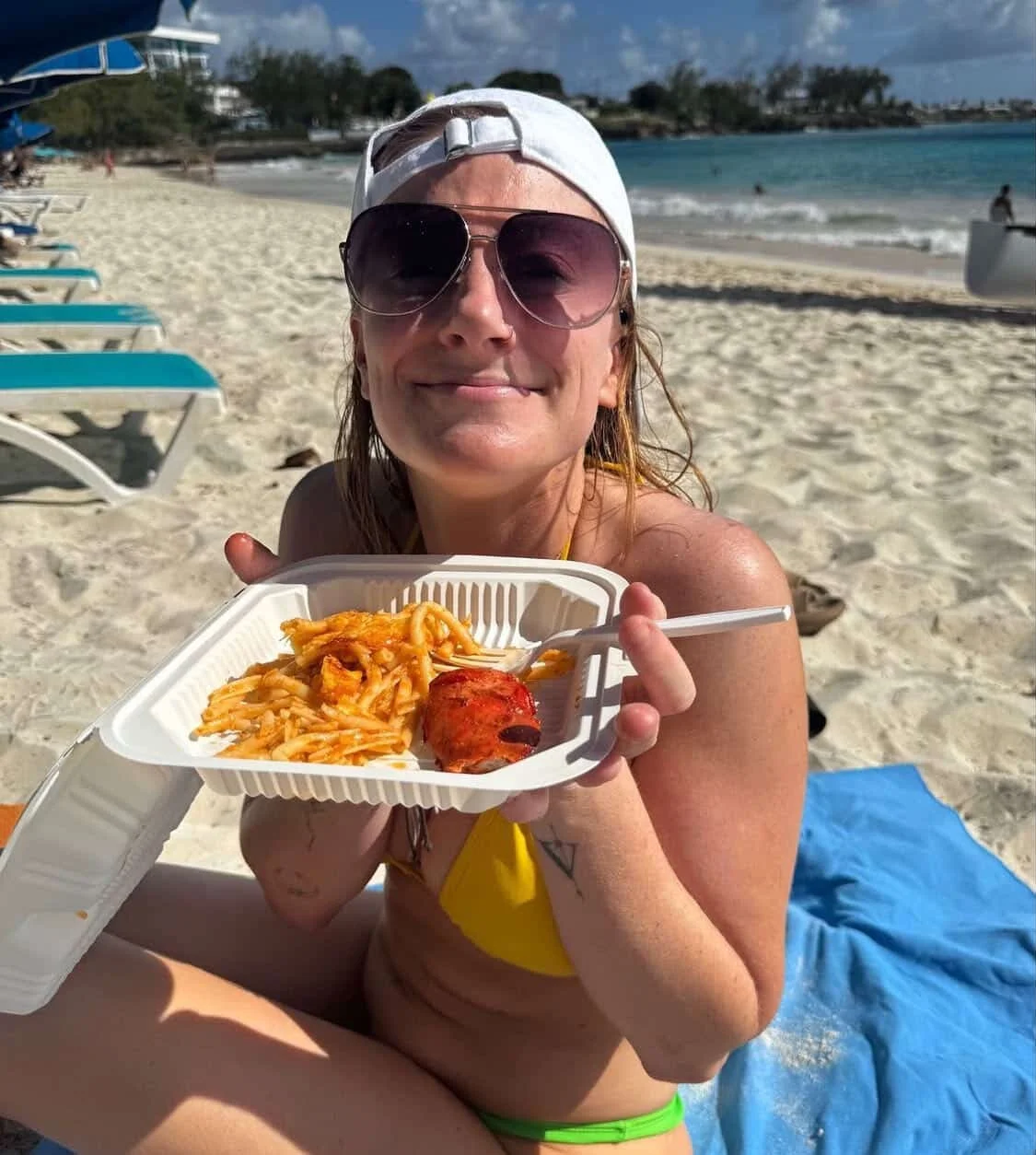 A woman sitting on a beach holding a takeout container with spaghetti and a meatball, smiling at the camera. She wears sunglasses, a white cap backwards, and a yellow bikini top with a blue towel beneath her.