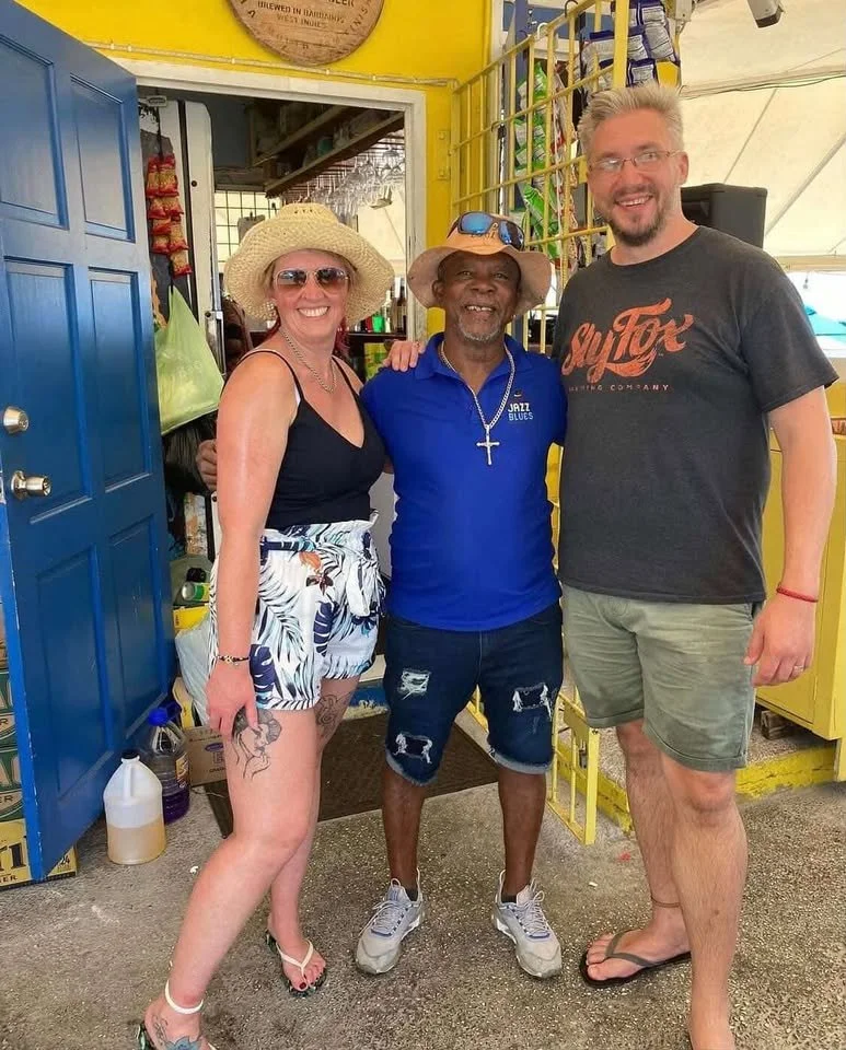 Three people smiling and standing close together inside a colorful shop or cafe. The woman on the left is wearing a straw hat, sunglasses, a black tank top, and patterned shorts. The man in the middle is wearing a blue polo shirt, a straw hat with sunglasses on top, and ripped shorts. The tall man on the right is wearing glasses, a black t-shirt with red writing, and green shorts. They are standing in front of a yellow and blue background, with various items visible around them.