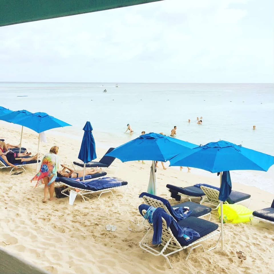 Beach scene with people swimming in the water, several blue umbrellas, and lounge chairs on the sandy shore.