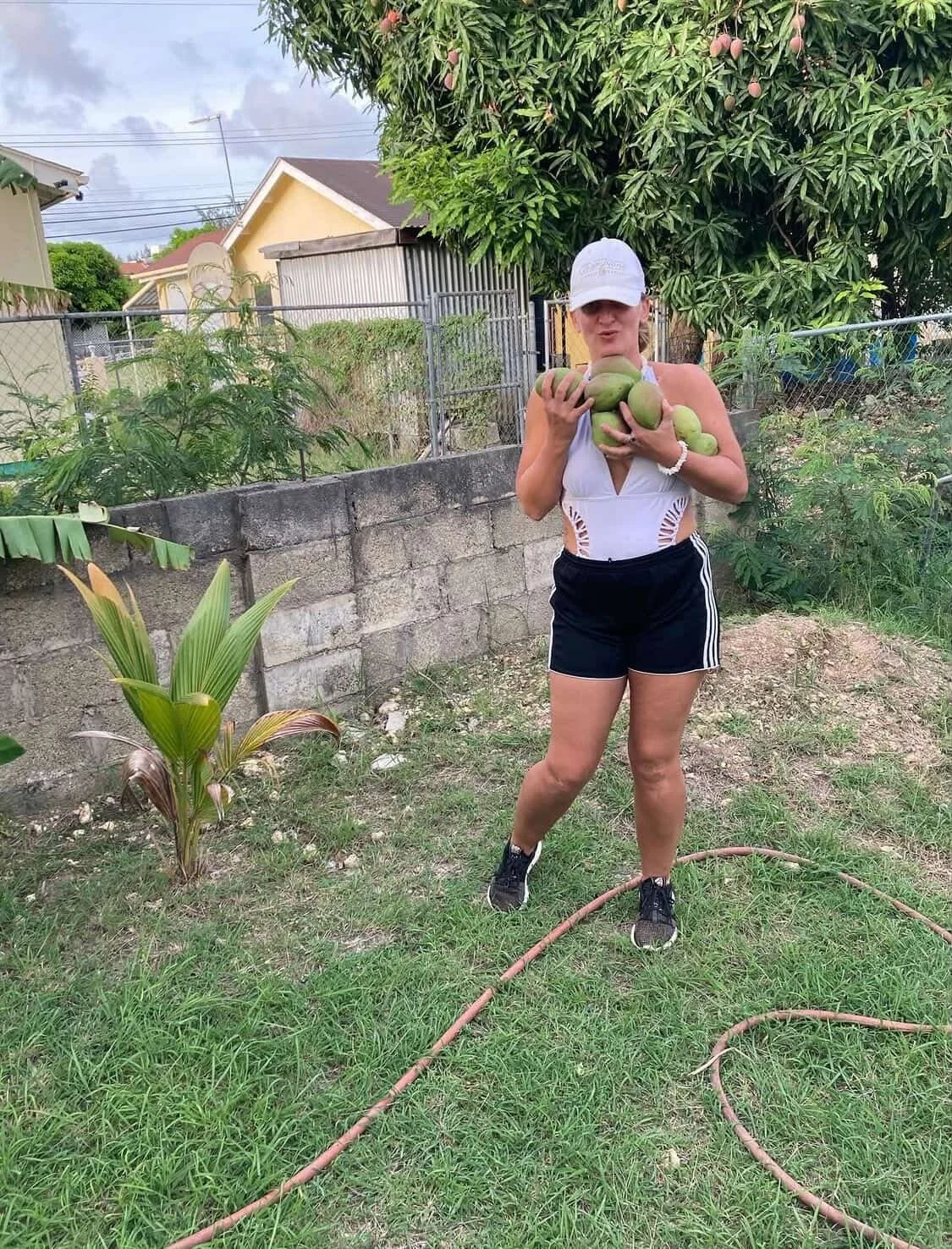 A woman standing in a backyard holding multiple green papayas, wearing a white cap, a white sleeveless top, black shorts, and black sneakers.