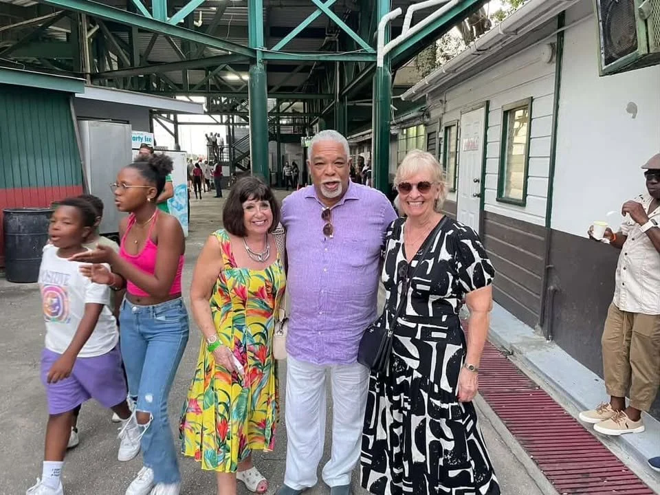 A group of four adults posing and smiling in an outdoor urban setting, with two women wearing colorful dresses and a man in a purple shirt, surrounded by children and other people in the background.