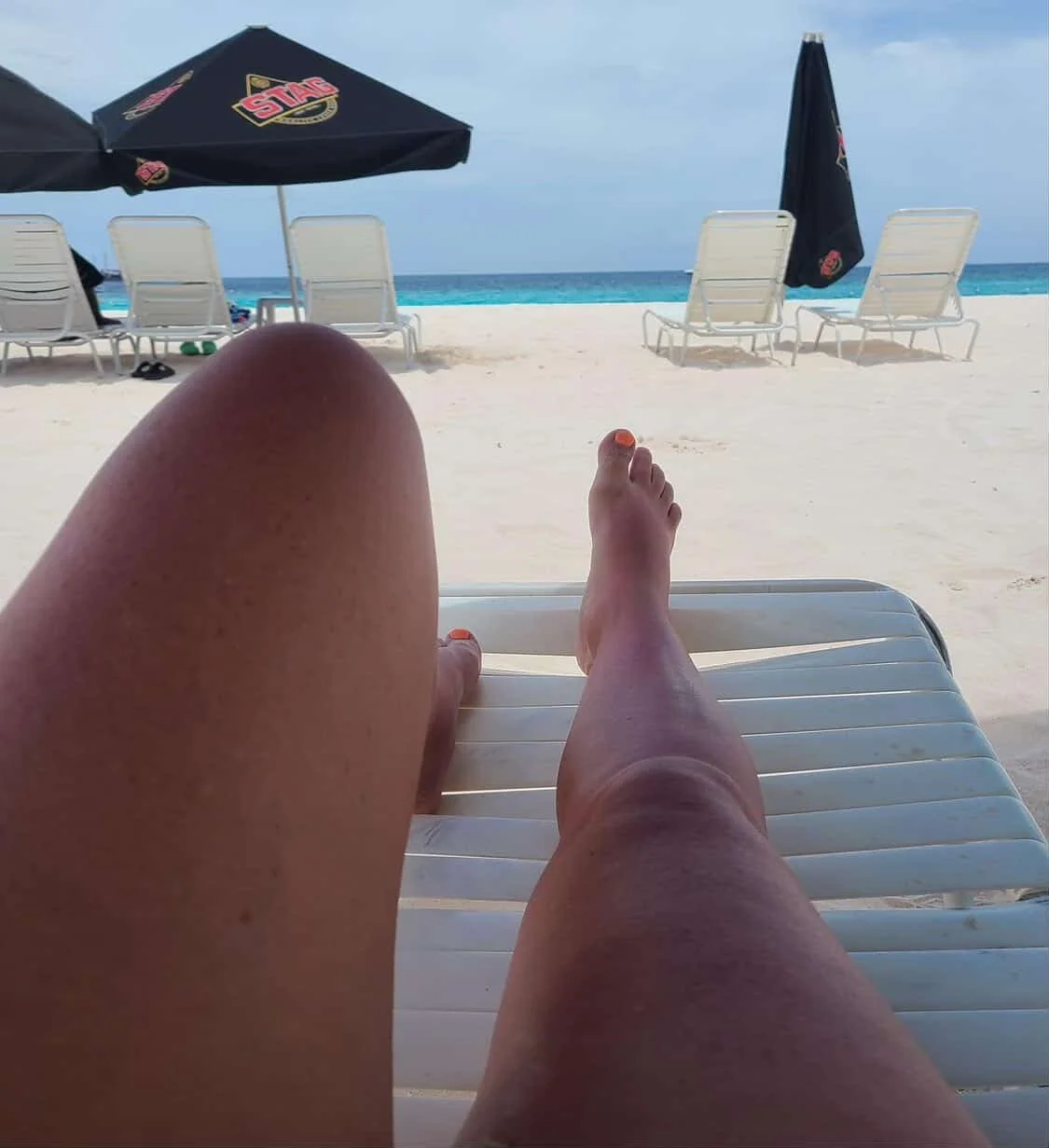 Person lounging on a white beach chair near the ocean, with feet in the foreground, beach umbrellas, and empty lounge chairs in the background, under a partly cloudy sky.