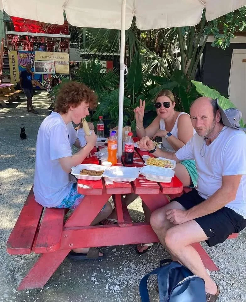 Three people sitting at a red outdoor picnic table with food bottles, eating and chatting, under an umbrella at a casual outdoor setting.