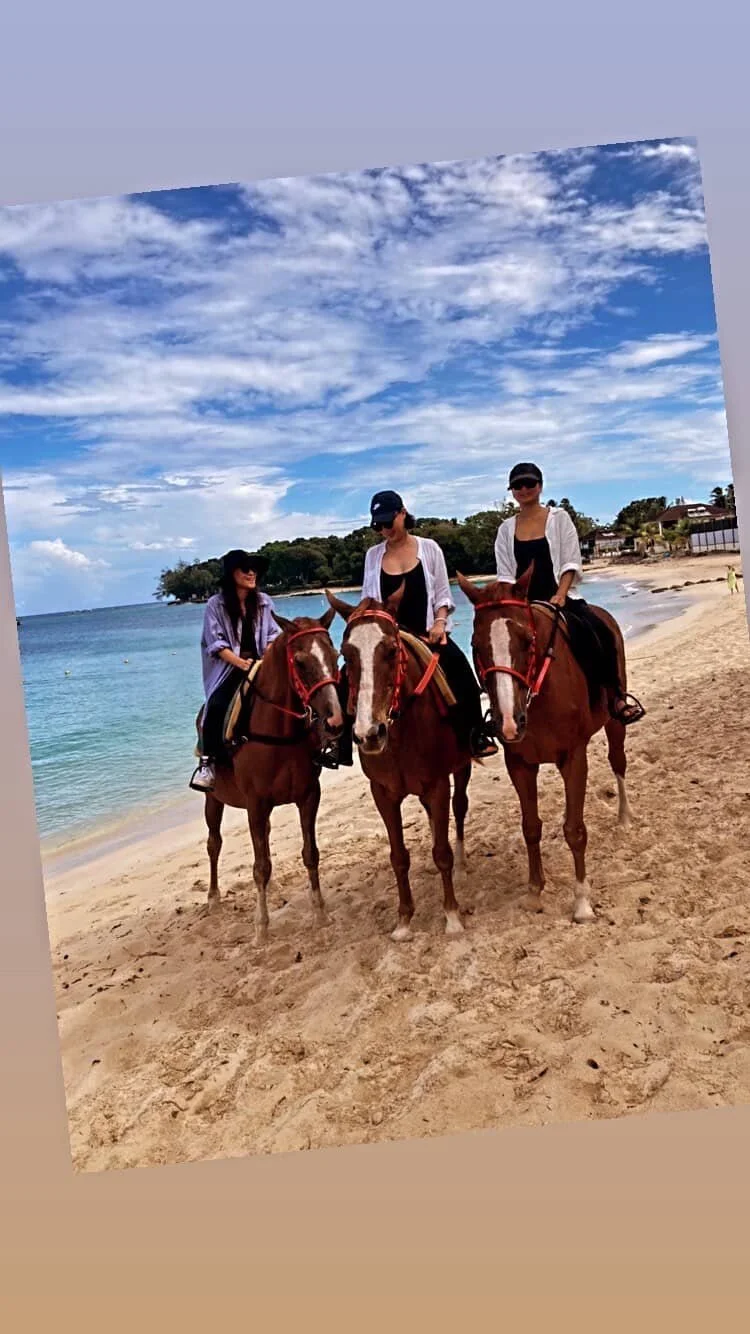 Three women riding horses on a sandy beach, with the ocean, blue sky with clouds, and some trees and buildings in the background.