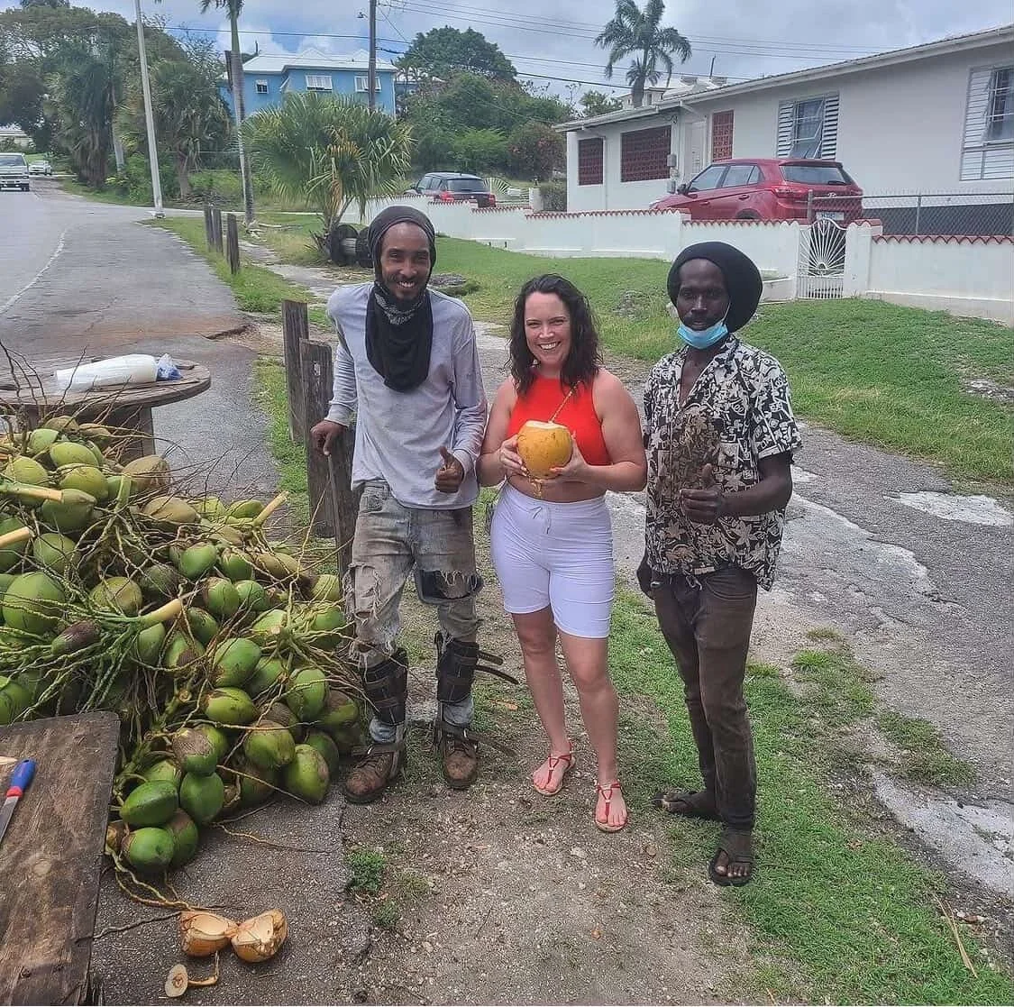 Three people standing outdoors on a street, smiling and giving thumbs up. One person holds a yellow coconut. There is a pile of coconuts on the ground beside them. The background shows a house, cars, and palm trees.
