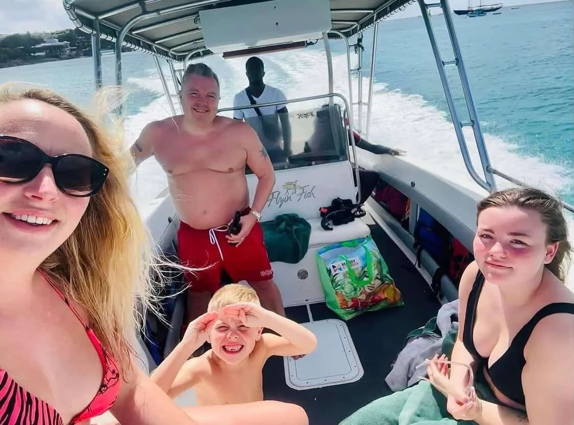 Group of five people on a boat enjoying a sunny day. Two women, a man, and two children are visible, with water and shoreline in the background.