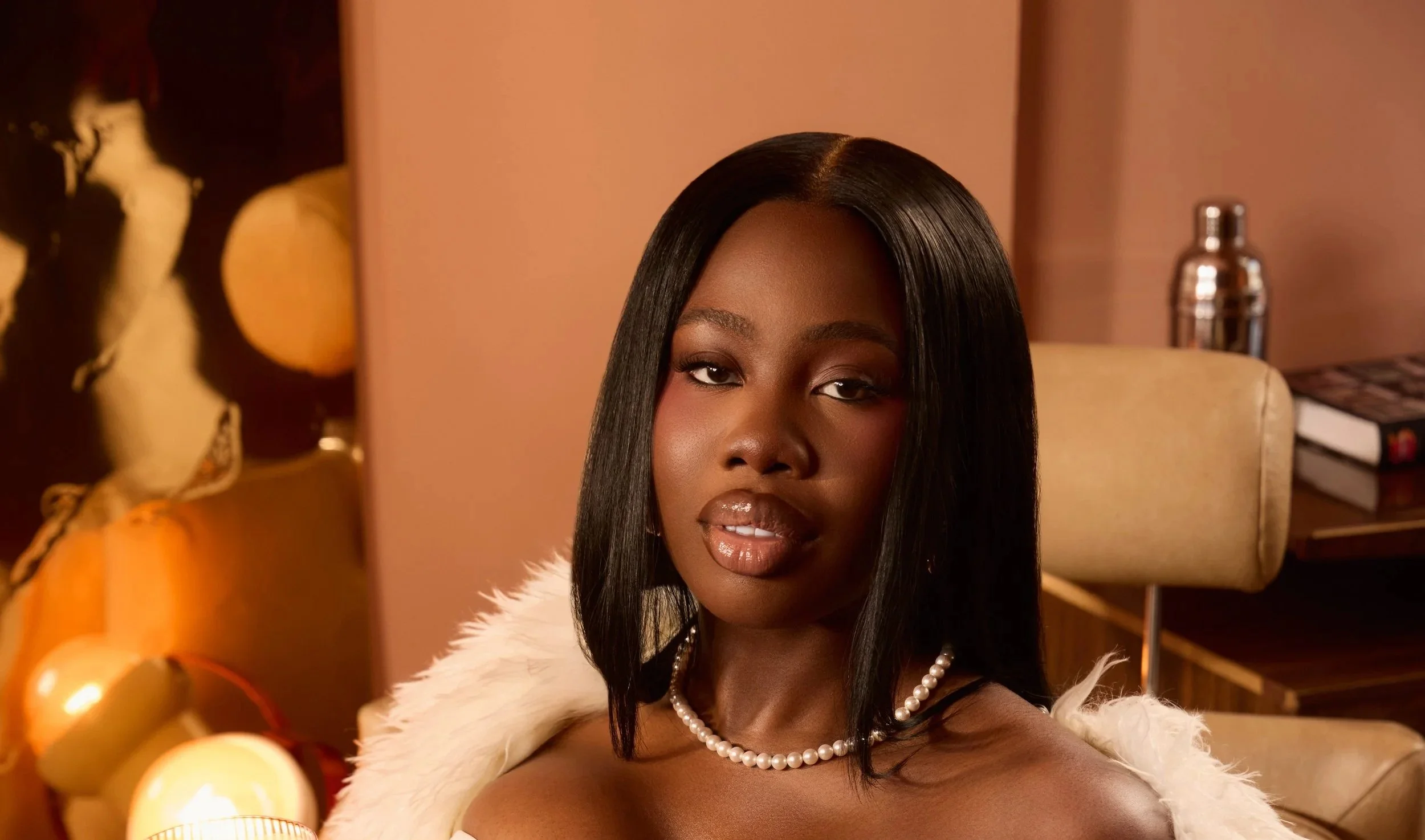 A woman with straight black hair, wearing a pearl necklace, a white fur stole, and makeup, is seated indoors with warm lighting. The background features orange pumpkins, Christmas ornaments, a chair, a table, and books.