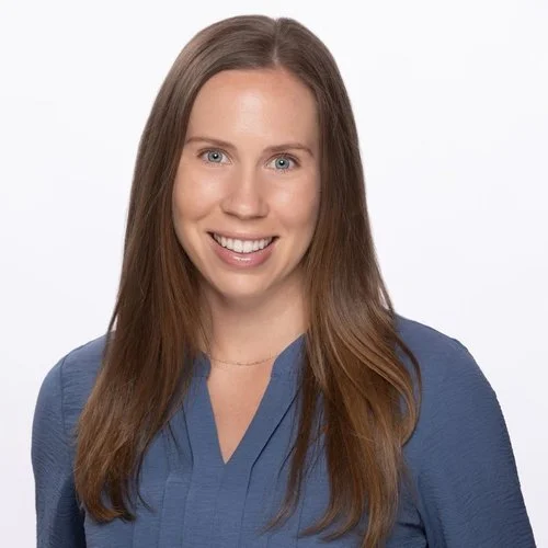 A woman with long brown hair, wearing a blue blouse, smiling against a plain white background.