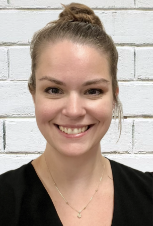 A young woman with fair skin and brown hair tied in a bun, smiling in front of a white brick wall.