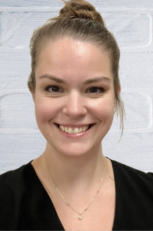 A young woman smiling with her hair in a bun, wearing a black top and a gold necklace, in front of a light gray wooden background.