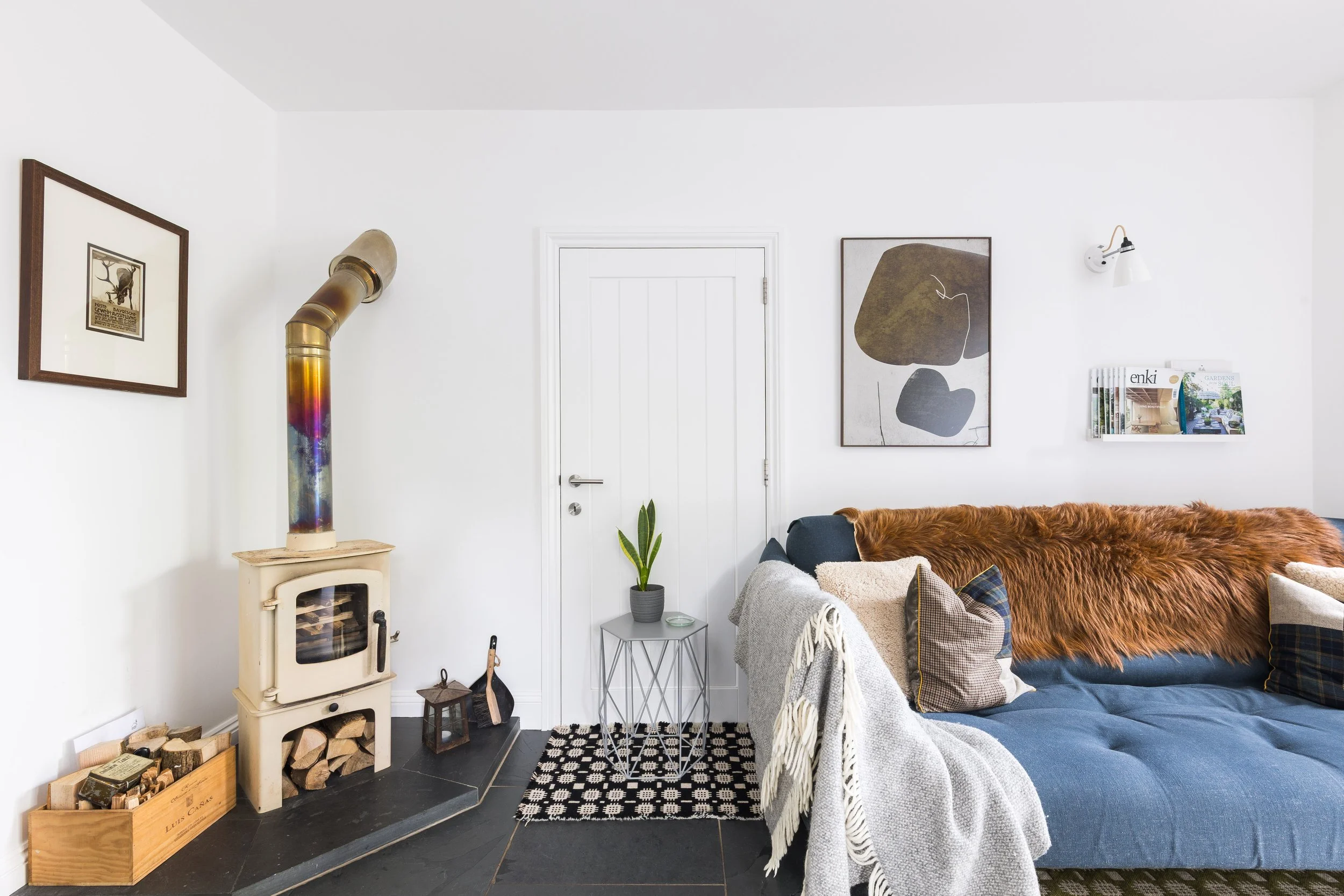 Living room with white walls, black tile floor, a white wood stove, logs stored in a wooden box, framed artwork, a window, a dark couch with assorted pillows, a cozy blanket, a small metal side table with a potted plant, and a modern wall sconce.