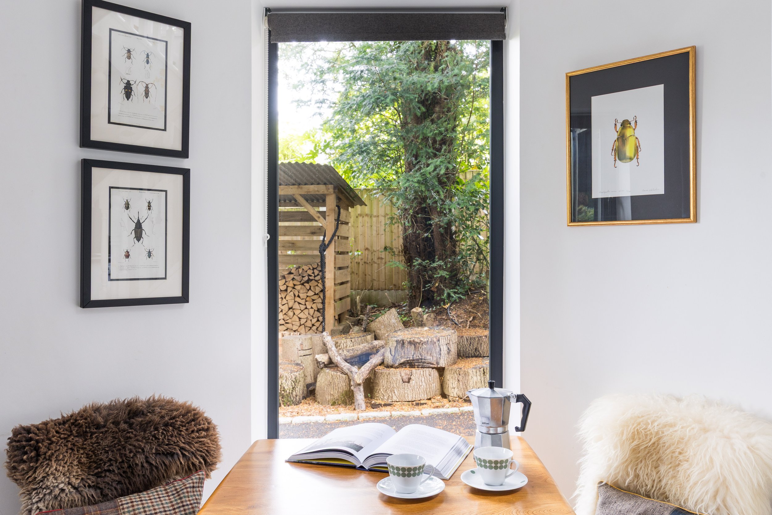 A cozy kitchen or dining area with a wooden table set with open books, cups, and a moka pot, decorated with fluffy pillows and fur throws on chairs. The background shows a large window with a view of a yard with a stack of firewood, trees, and a wood