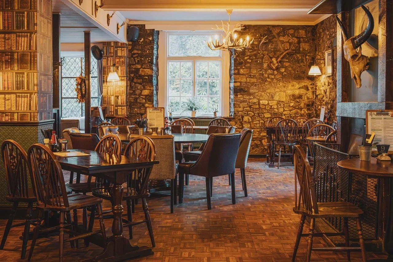 A cozy restaurant with wooden tables and chairs, a stone wall, large window, and decorative animal skulls and antlers