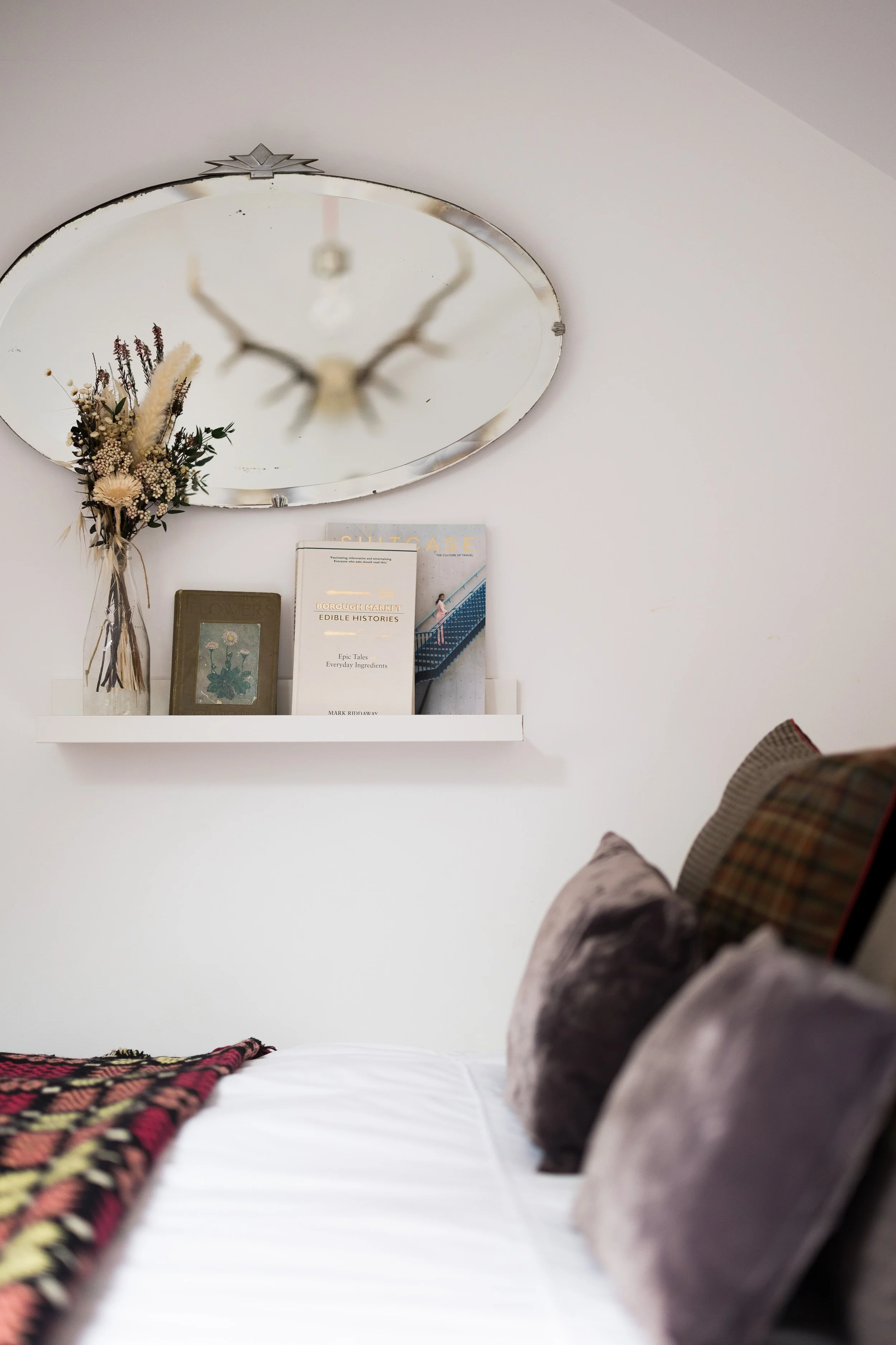 A bedroom with a white bed and a shelf on the wall. The shelf has books, a framed picture, and dried flowers in a vase. The bed has a blanket and pillows. There is a mirror on the ceiling reflecting part of the room.