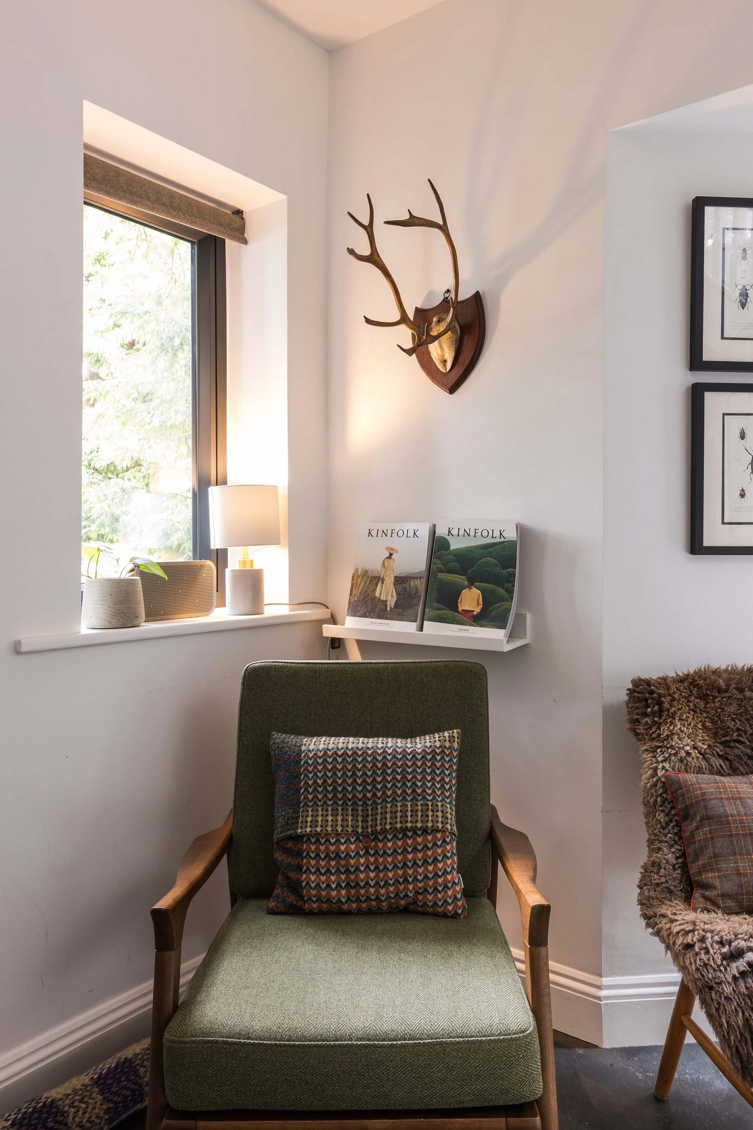 A cozy reading nook with a green armchair and matching footstool, decorated with a colorful cushion, placed near a window with a lamp and decor items on the sill, and framed artwork on the wall.