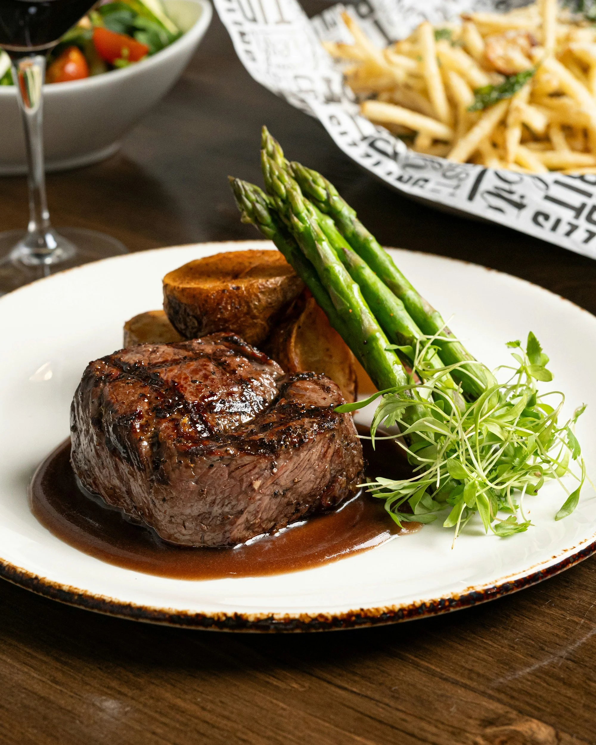 A plate with a cooked beef steak topped with a brown sauce, garnished with fresh green asparagus and microgreens. In the background, there is a bowl of salad with greens and cherry tomatoes, and a serving of French fries on parchment paper.