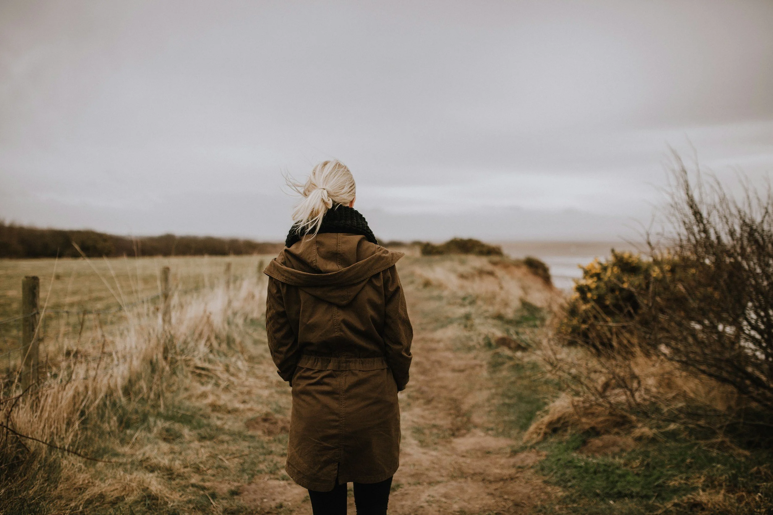 A woman with blonde hair tied in a ponytail, wearing a brown coat and black scarf, standing outdoors on a cloudy day near a sandy beach with sparse vegetation.