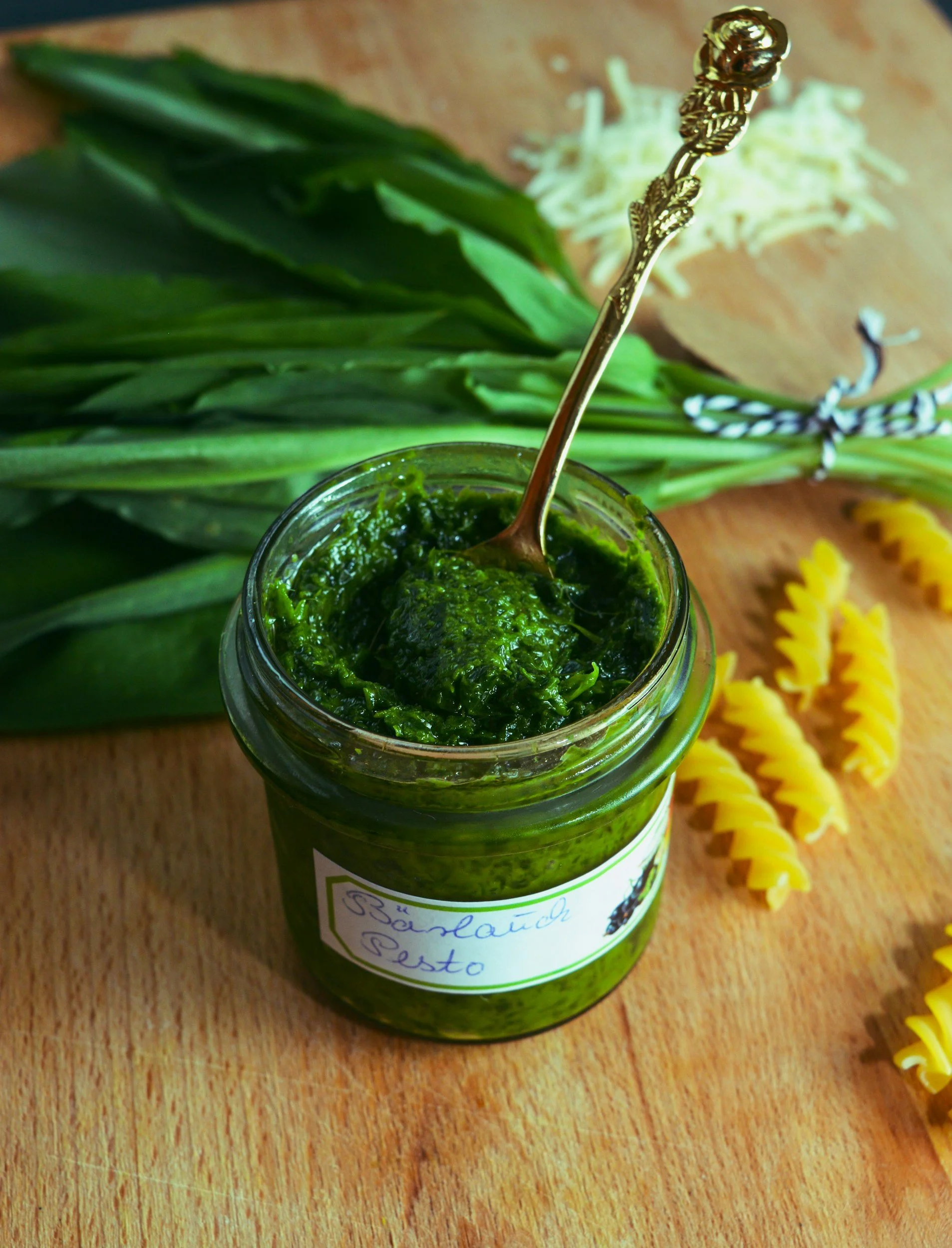 A jar of green pesto with a silver spoon inside, placed on a wooden surface next to a bunch of green onions and uncooked yellow pasta.