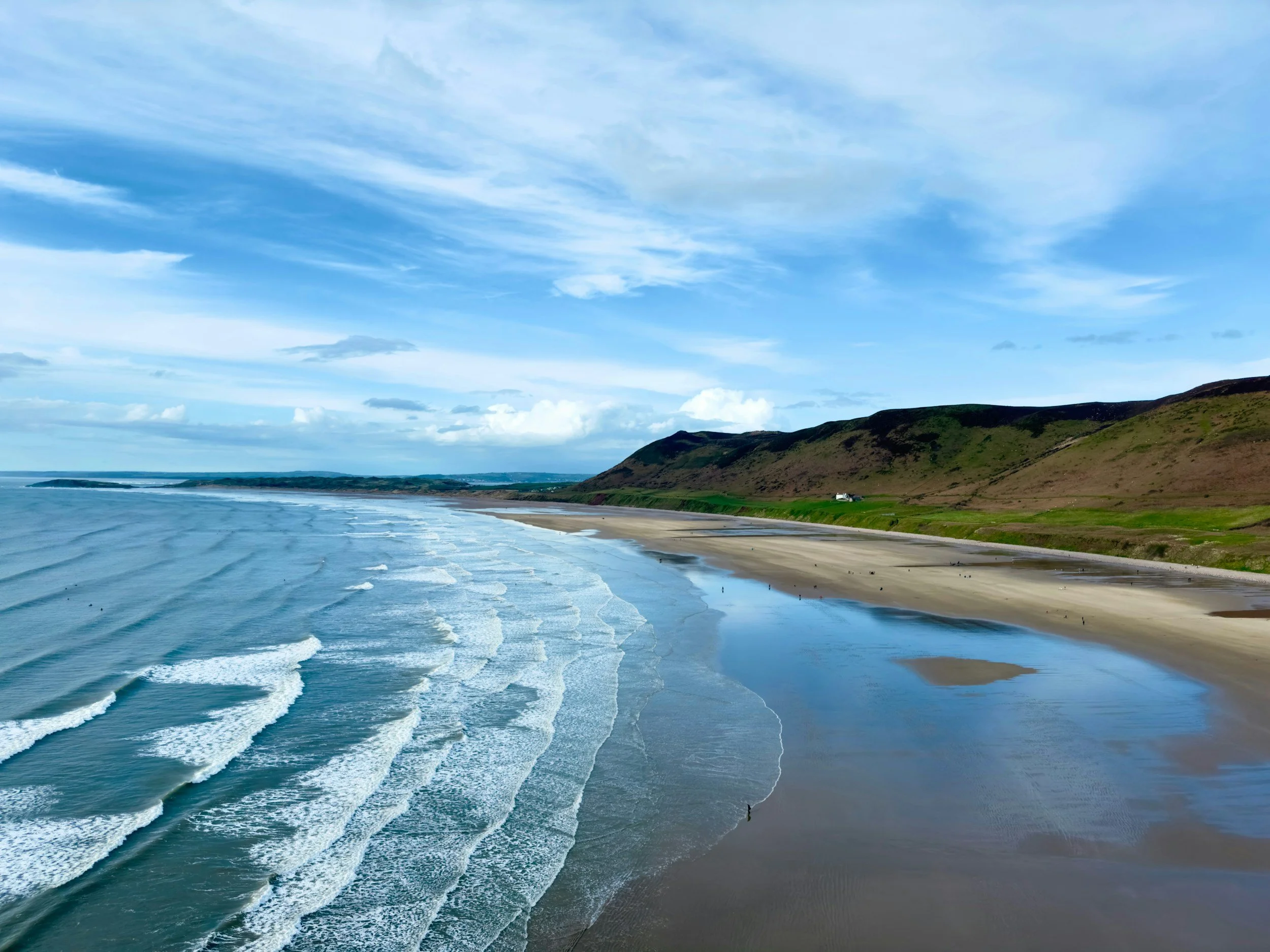 A scenic view of a beach with waves crashing onto the shore, green hills in the background, and a partly cloudy blue sky.