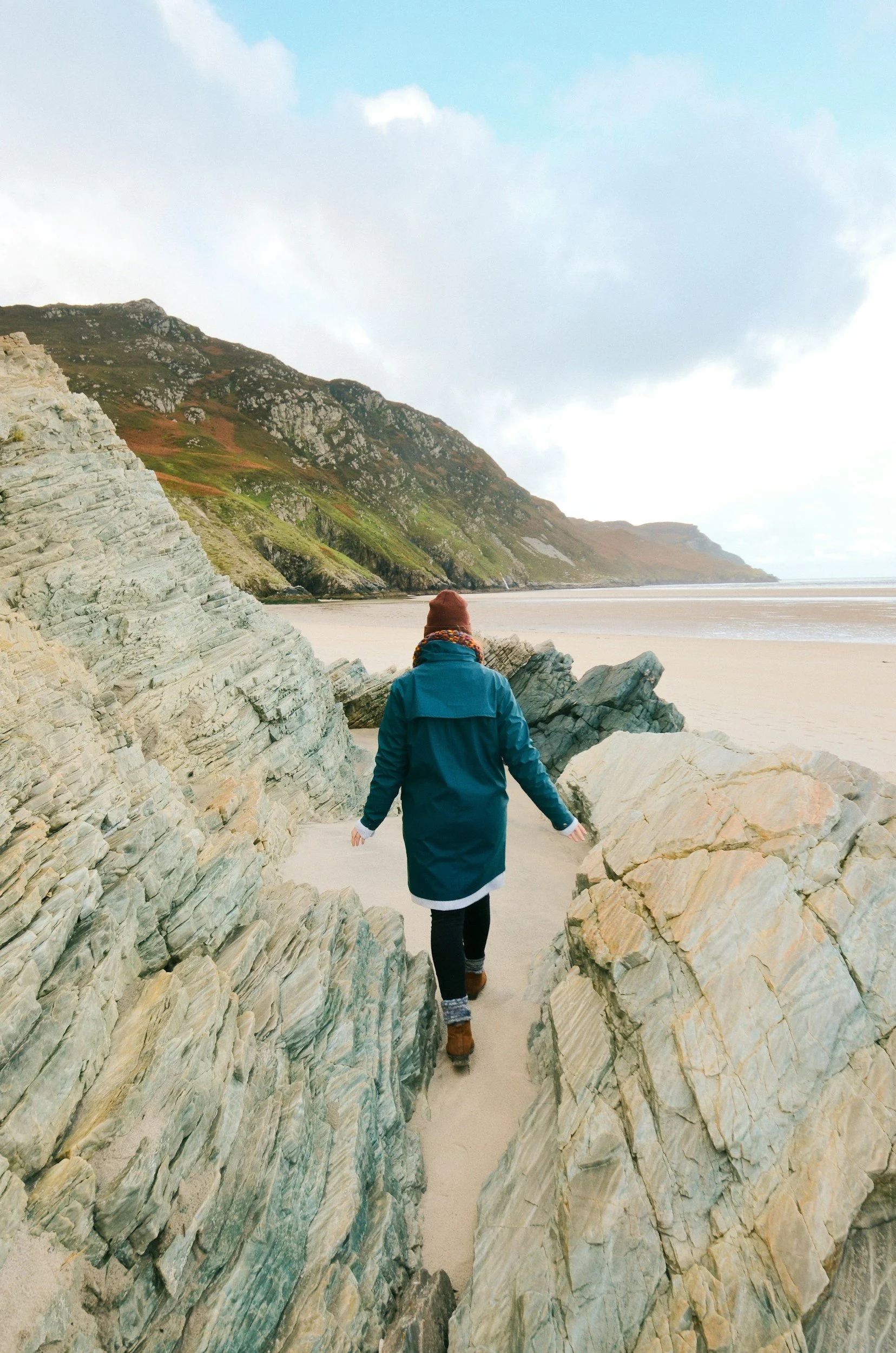 A woman walking through a narrow gap between large, layered rocks on a sandy beach with green hills and a cloudy sky in the background.