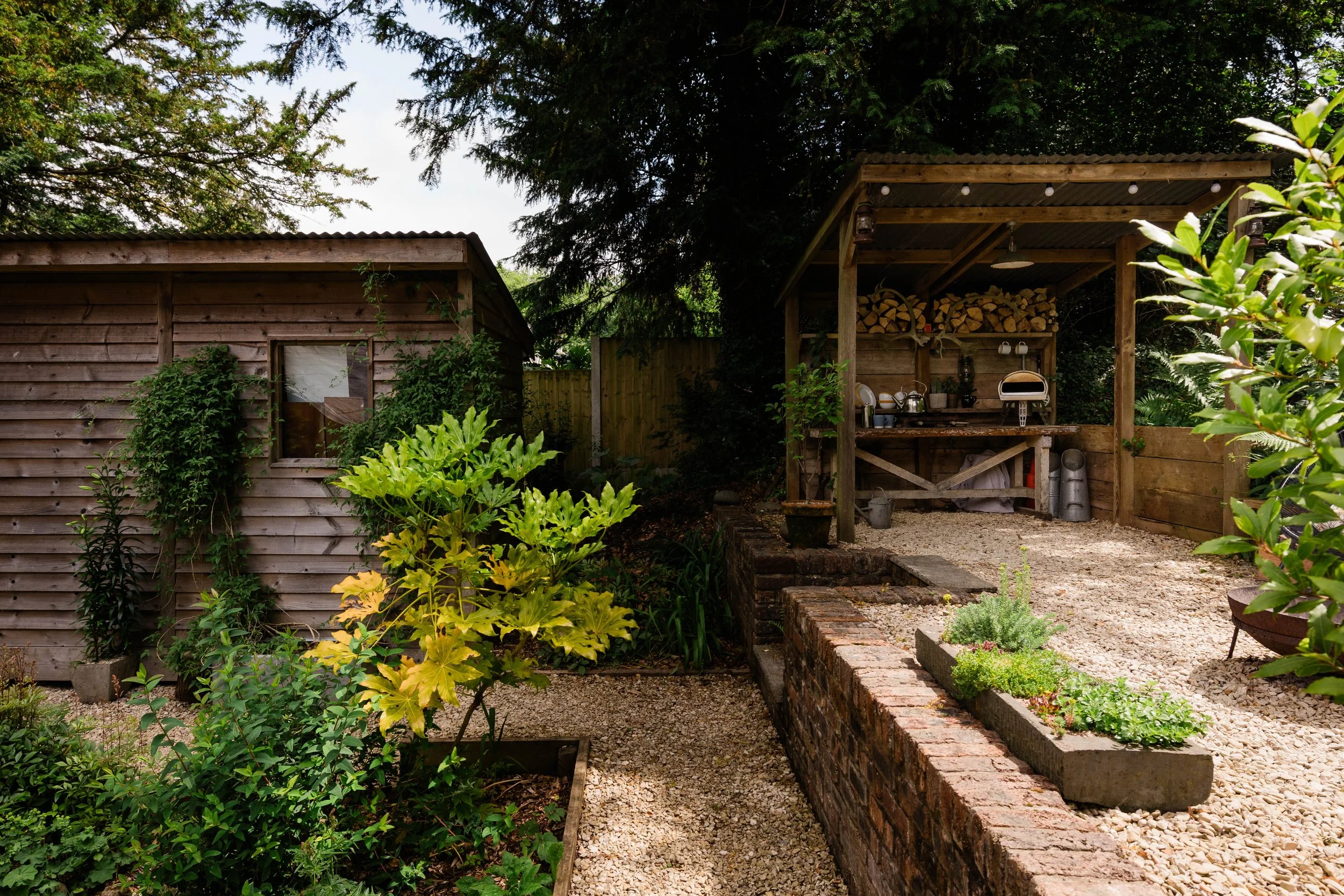 A backyard garden scene with a wooden shed and a wood-covered outdoor kitchen with storage for firewood. There are green plants and shrubs in the garden, and a gravel pathway runs through it.