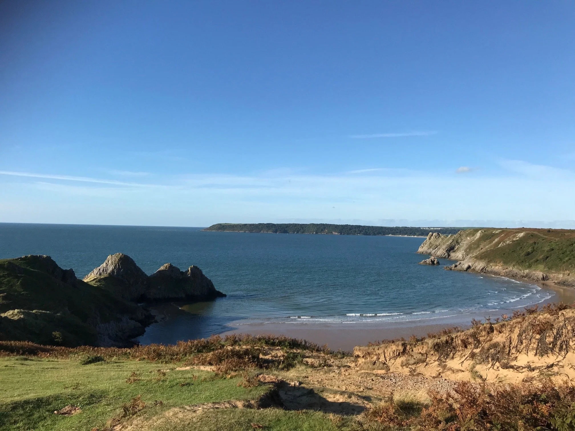 A coastal scene with rocky cliffs, a sandy beach, a body of water, and a hill in the background under a clear blue sky.