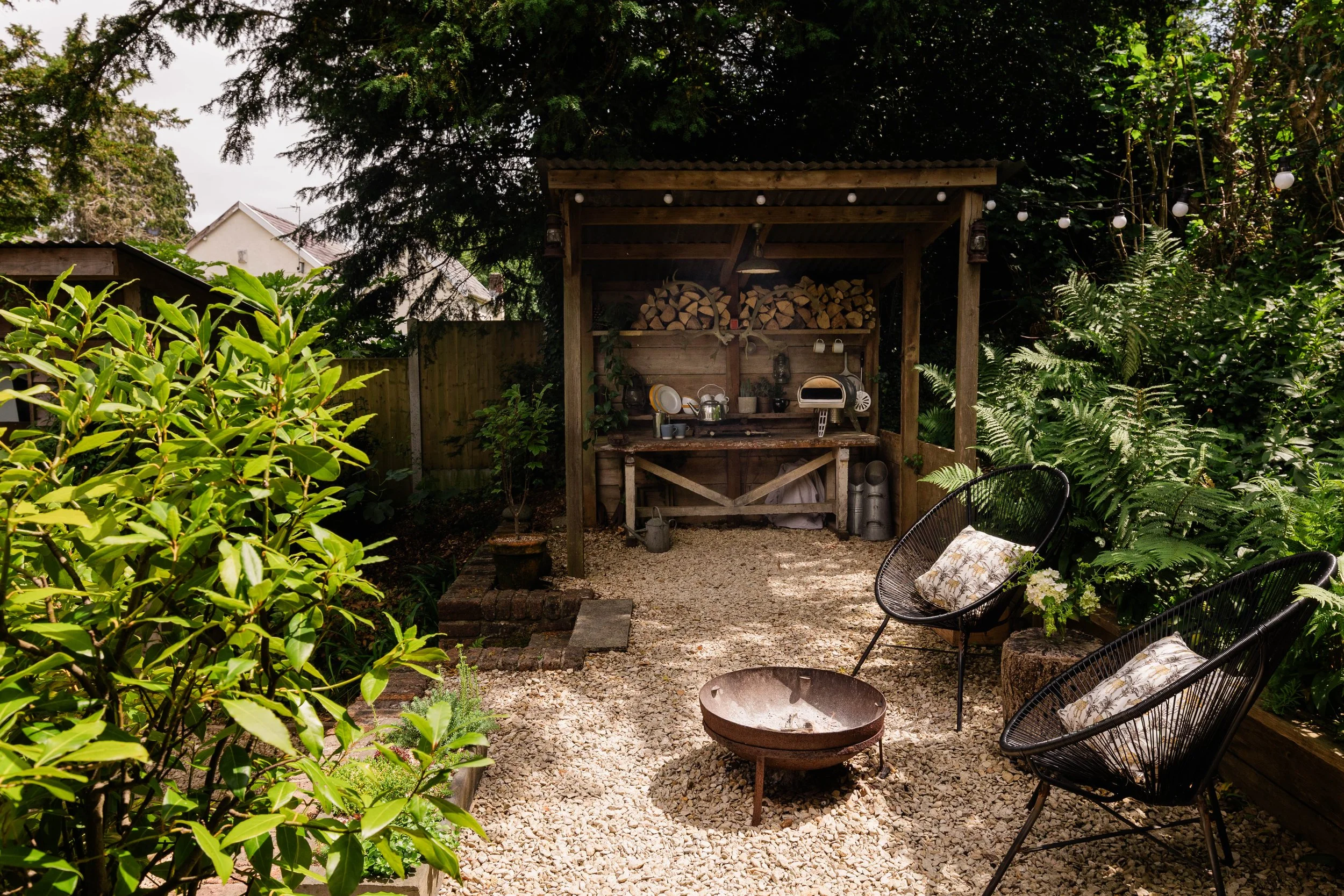 A cozy backyard outdoor space with a wood fire pit, black wire chairs with floral cushions, a wooden shed with firewood, plants, and string lights under a large tree.