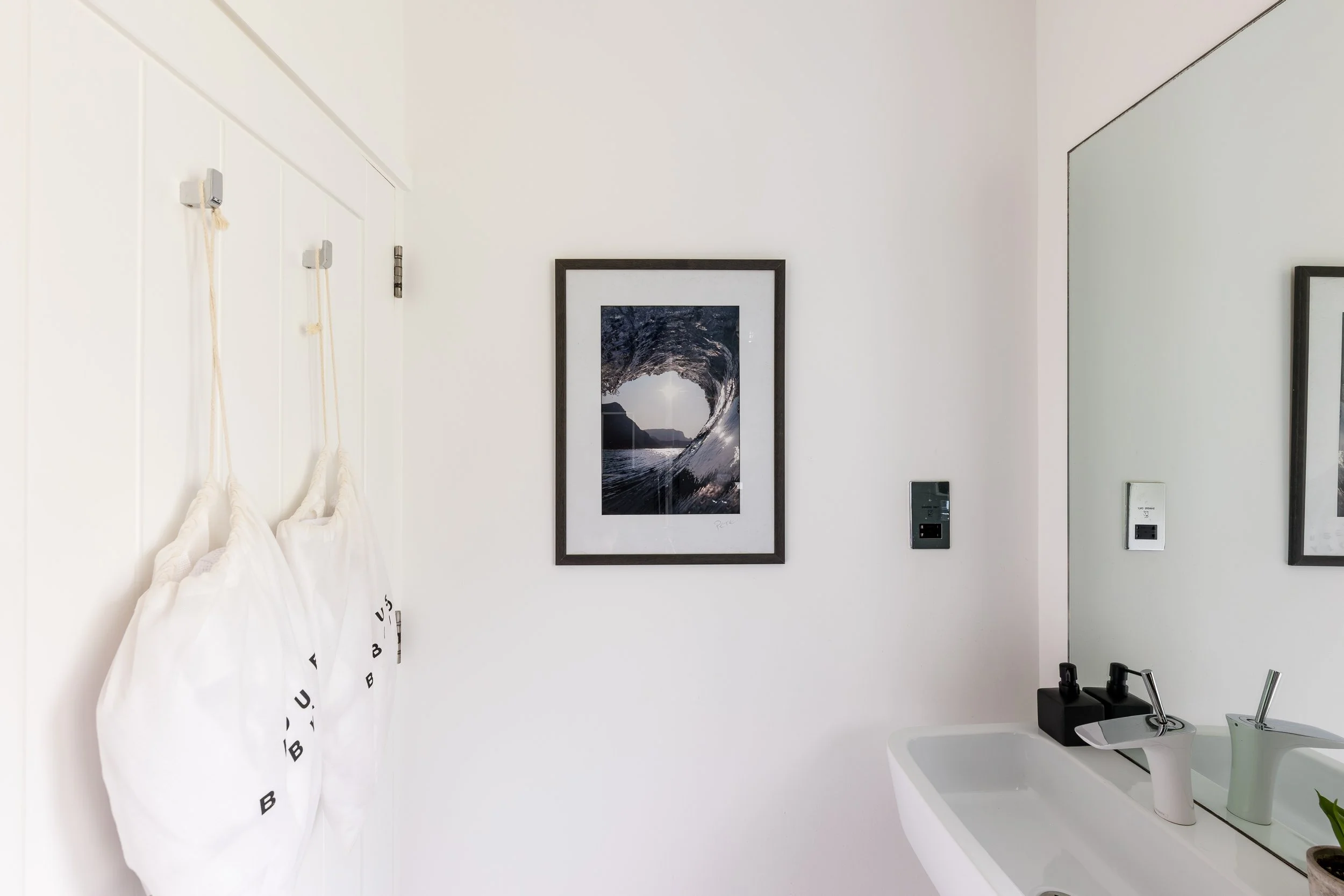 A modern bathroom with white walls, a framed ocean photograph, and a large mirror. A white towel is hanging on the wall to the left and is printed with black letters. There is a small sink with a soap dispenser and other toiletries on the right.
